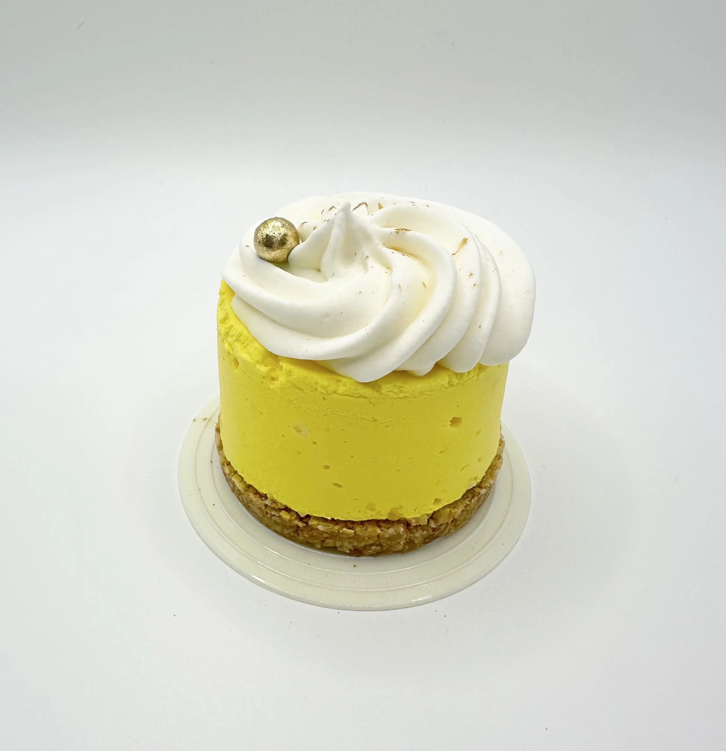 Yellow cupcake with white frosting swirl topped with a small gold ball and gold accents, placed on a clear white circular dish against a white background.