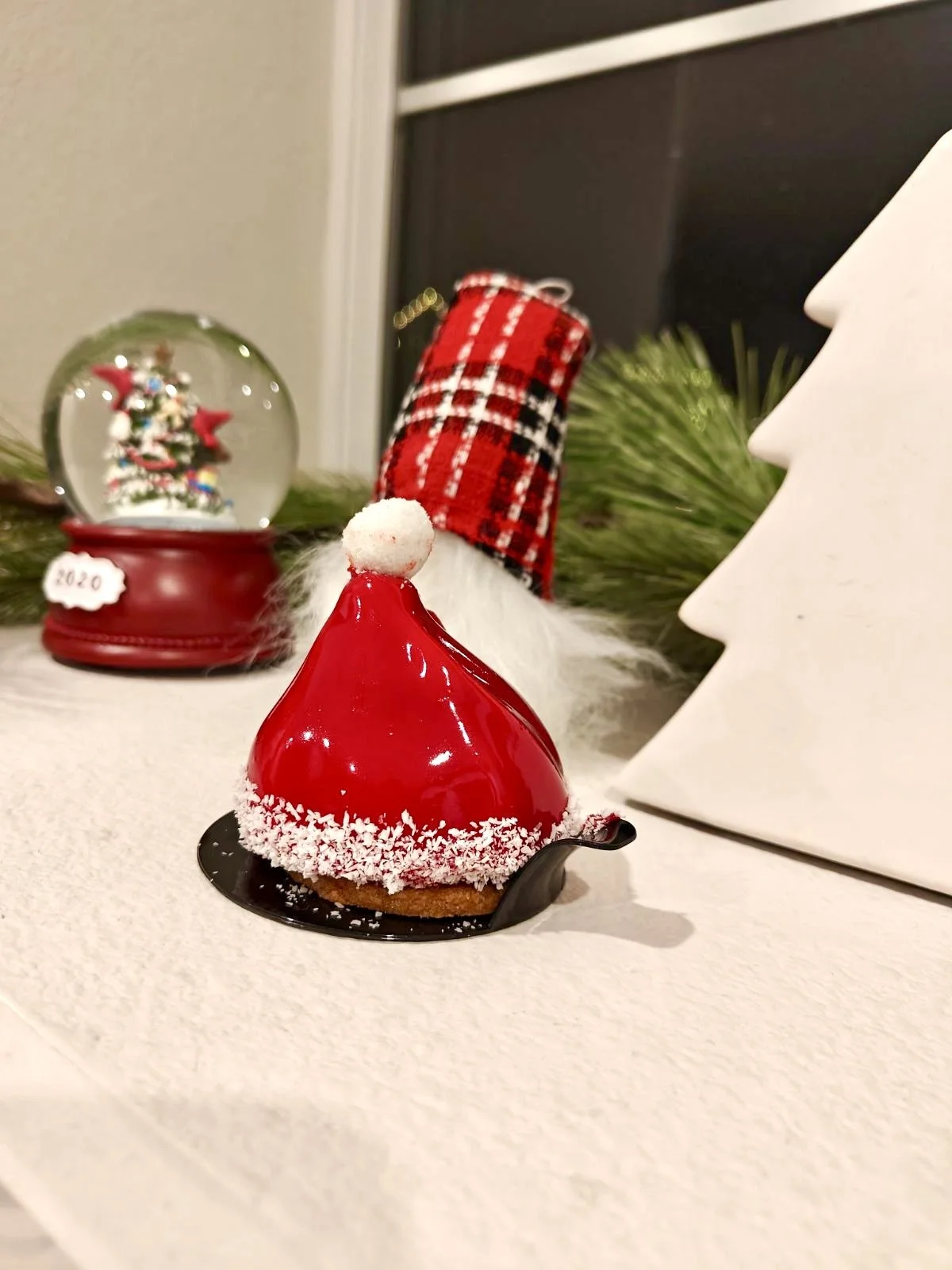 Christmas-themed dessert with red glaze and white decorations, surrounded by holiday decorations including a snow globe, a white Christmas tree sculpture, and a small Santa hat with a plaid pattern in the background.