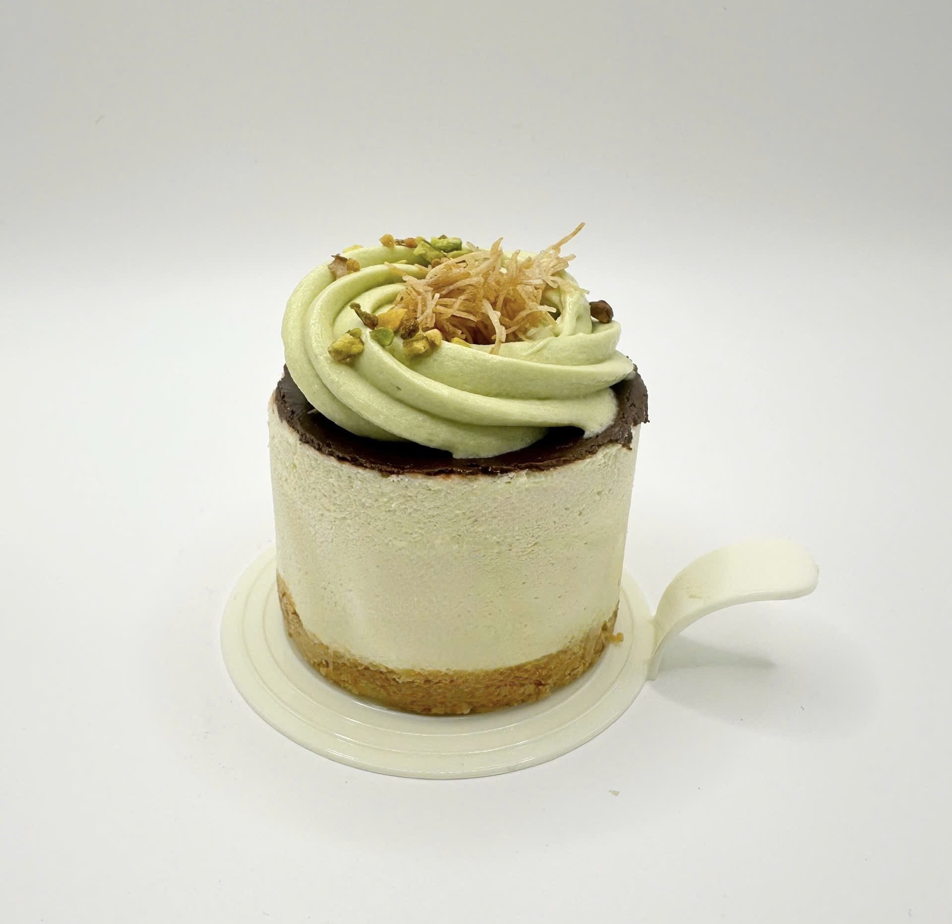 Close-up of a small dessert with white exterior, dark chocolate layer, light green frosting topping, topped with crushed pistachios and thin caramelized shredded ingredient, on a white plate against a plain background.