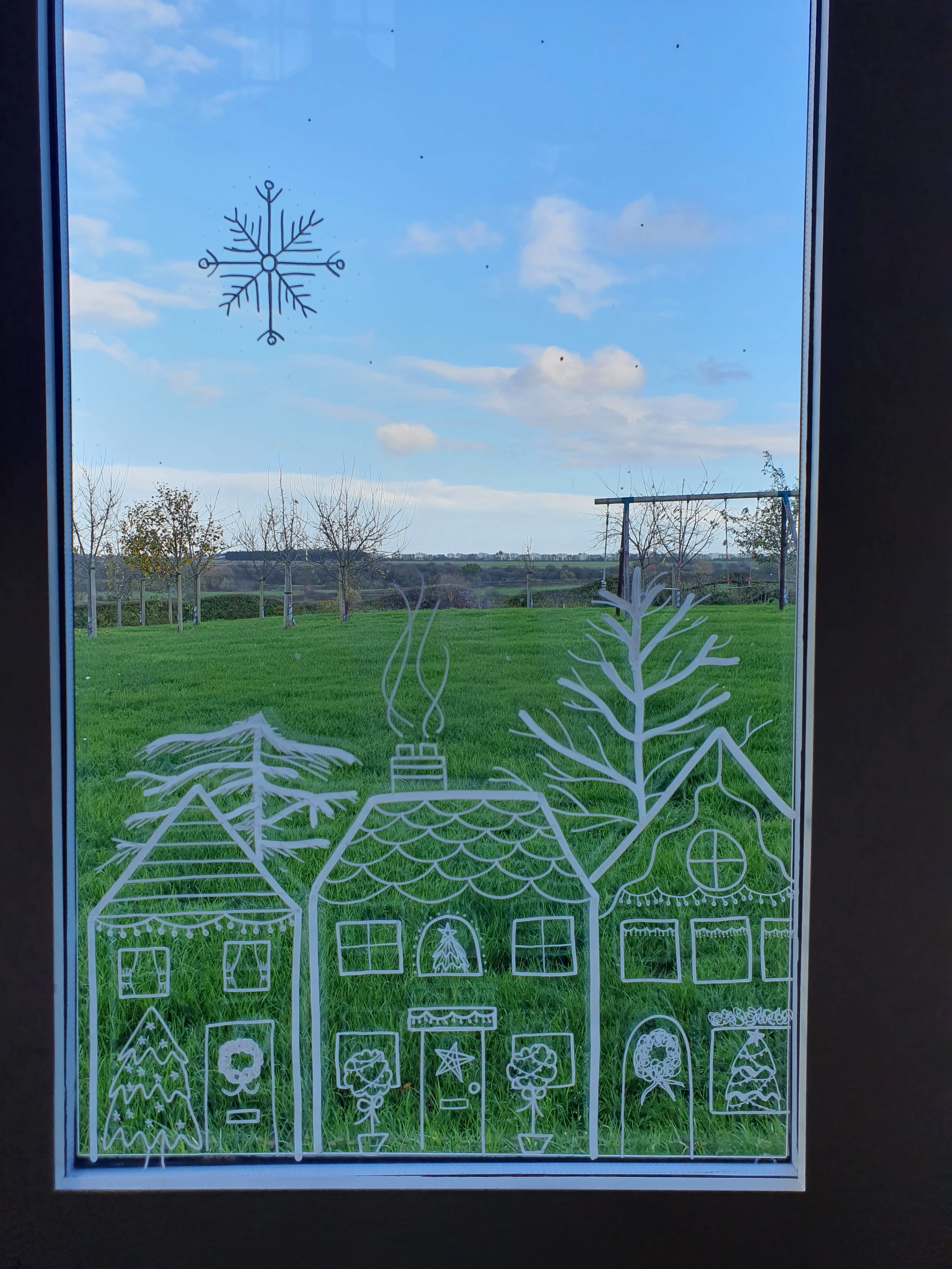 Window with white Christmas-themed drawings of houses and trees, against a background of green grass and a cloudy sky.