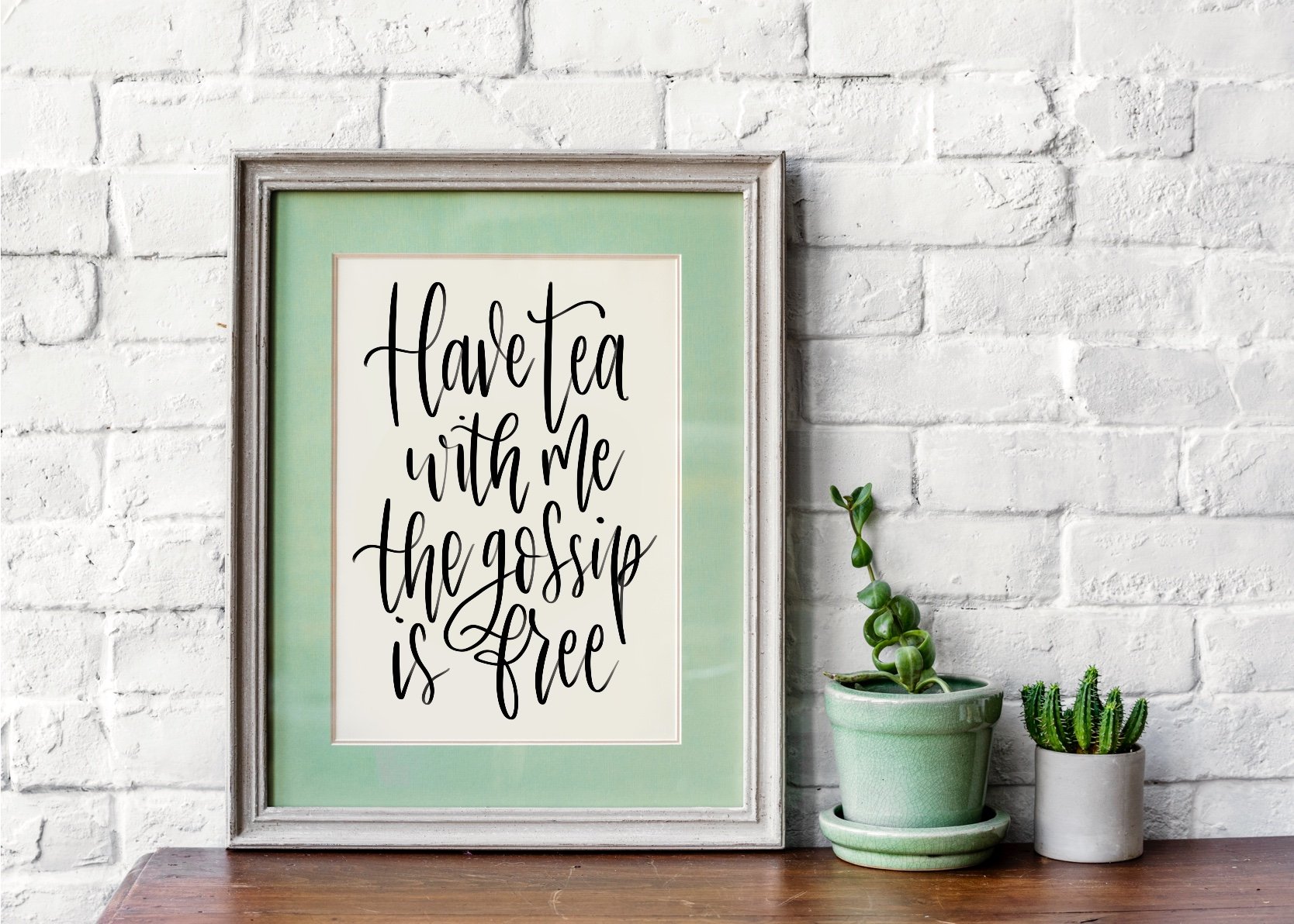 Framed quote saying "Have tea with me the gossip is free" on a wooden table with potted plants against a white brick wall.