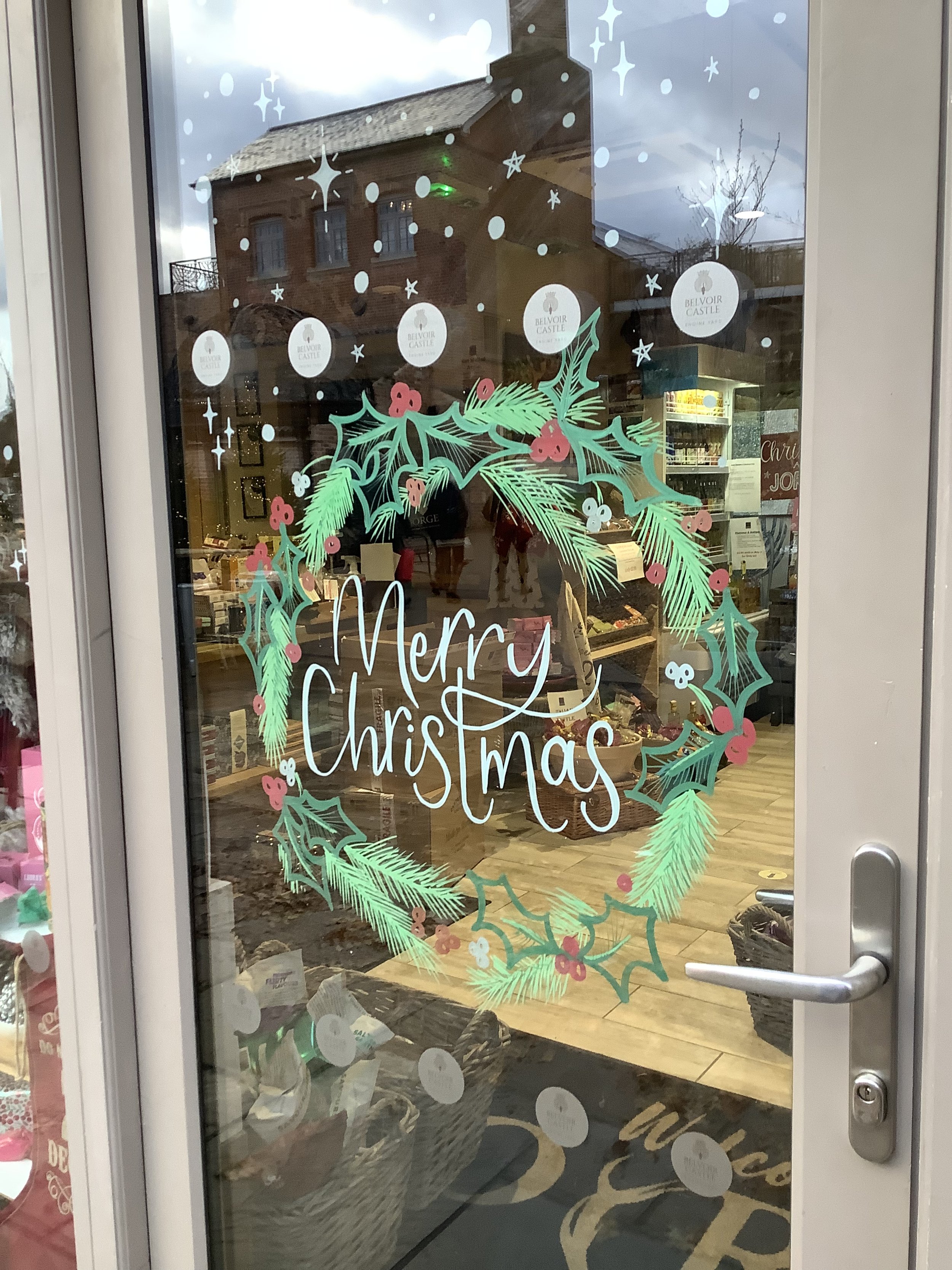 Glass door with a "Merry Christmas" wreath decoration, decorated with festive pine leaves, holly, and berries, reflecting a brick building.
