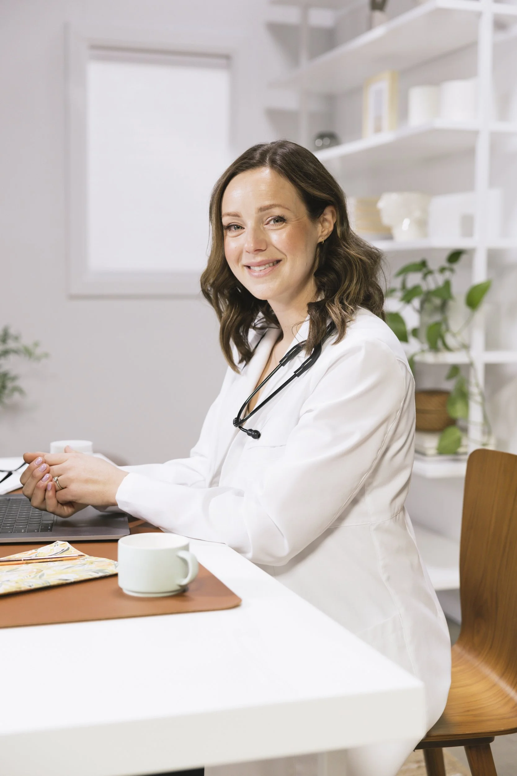 Dr. Miranda Naylor in white lab coat with stethoscope around neck sitting at desk