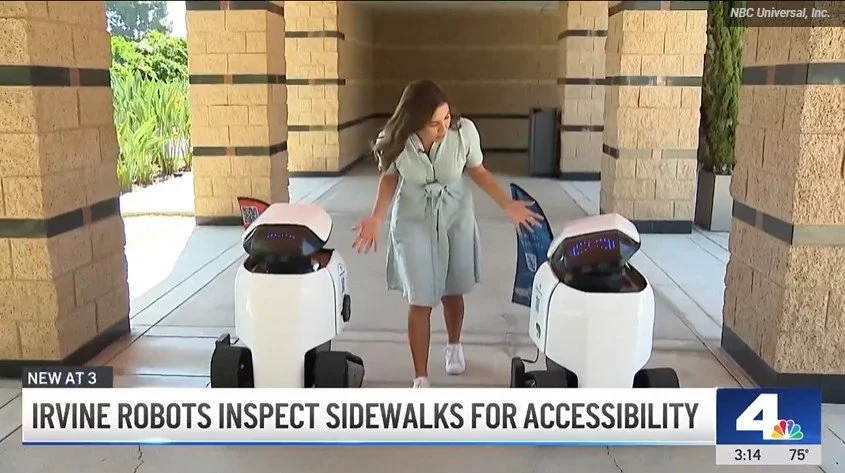 Two Dax robots walk with Amber Frias while an NBC crew films in Irvine.