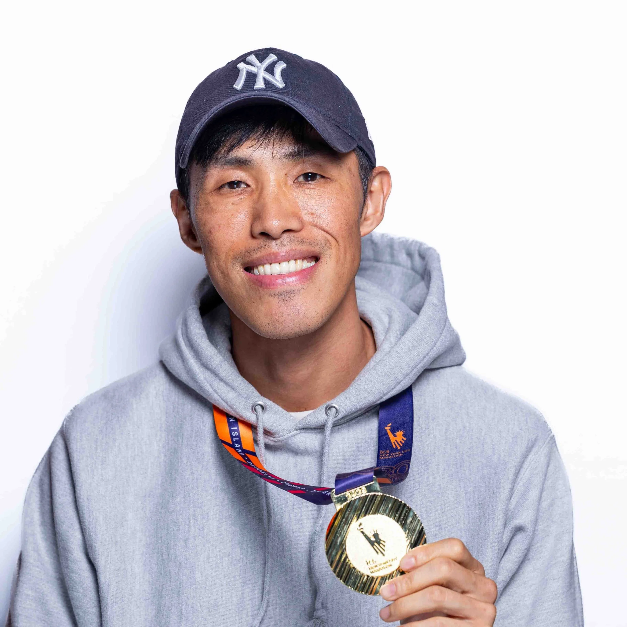Man wearing a New York Yankees cap and gray hoodie, holding a gold medal with a blue ribbon, smiling at the camera.