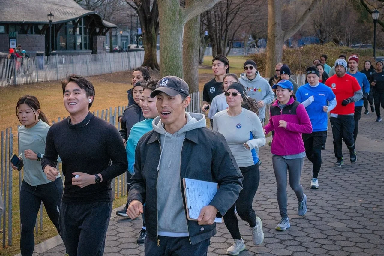 A group of diverse people participating in a group run in a park during a cool or mild day, wearing athletic clothing and some with headphones or clipboards.