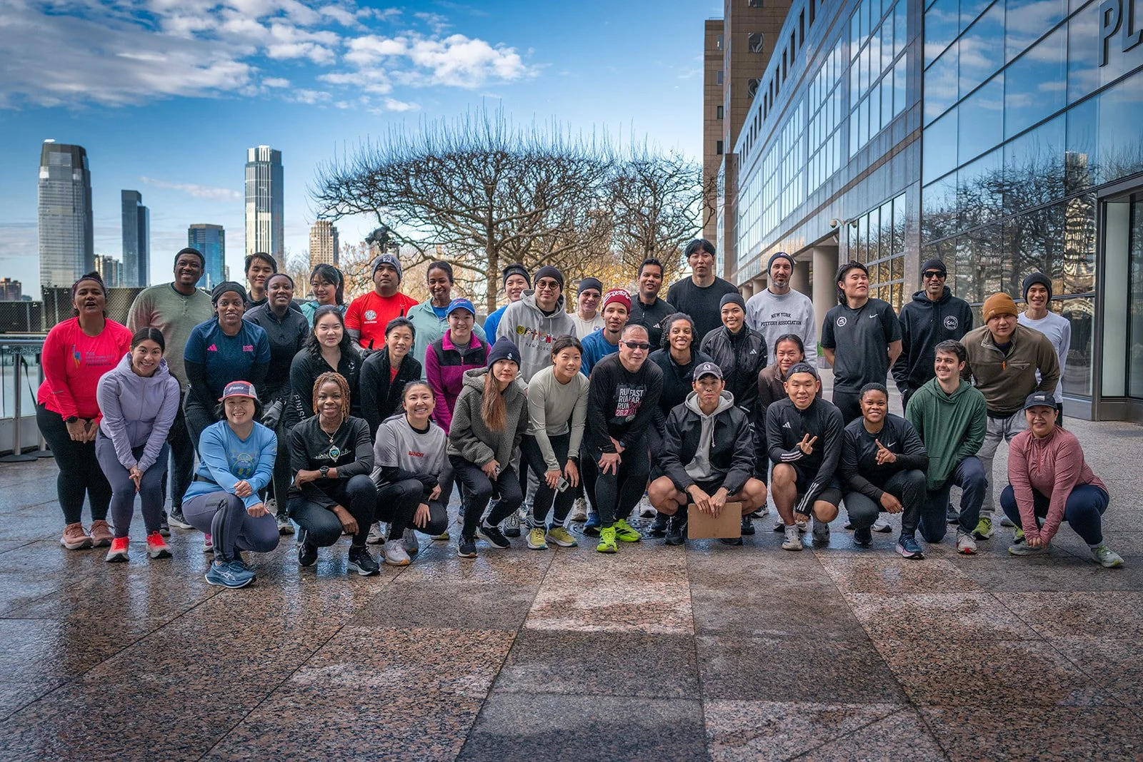 Group of diverse people in athletic clothing gathered outdoors in an urban setting with skyscrapers and leafless trees in the background.