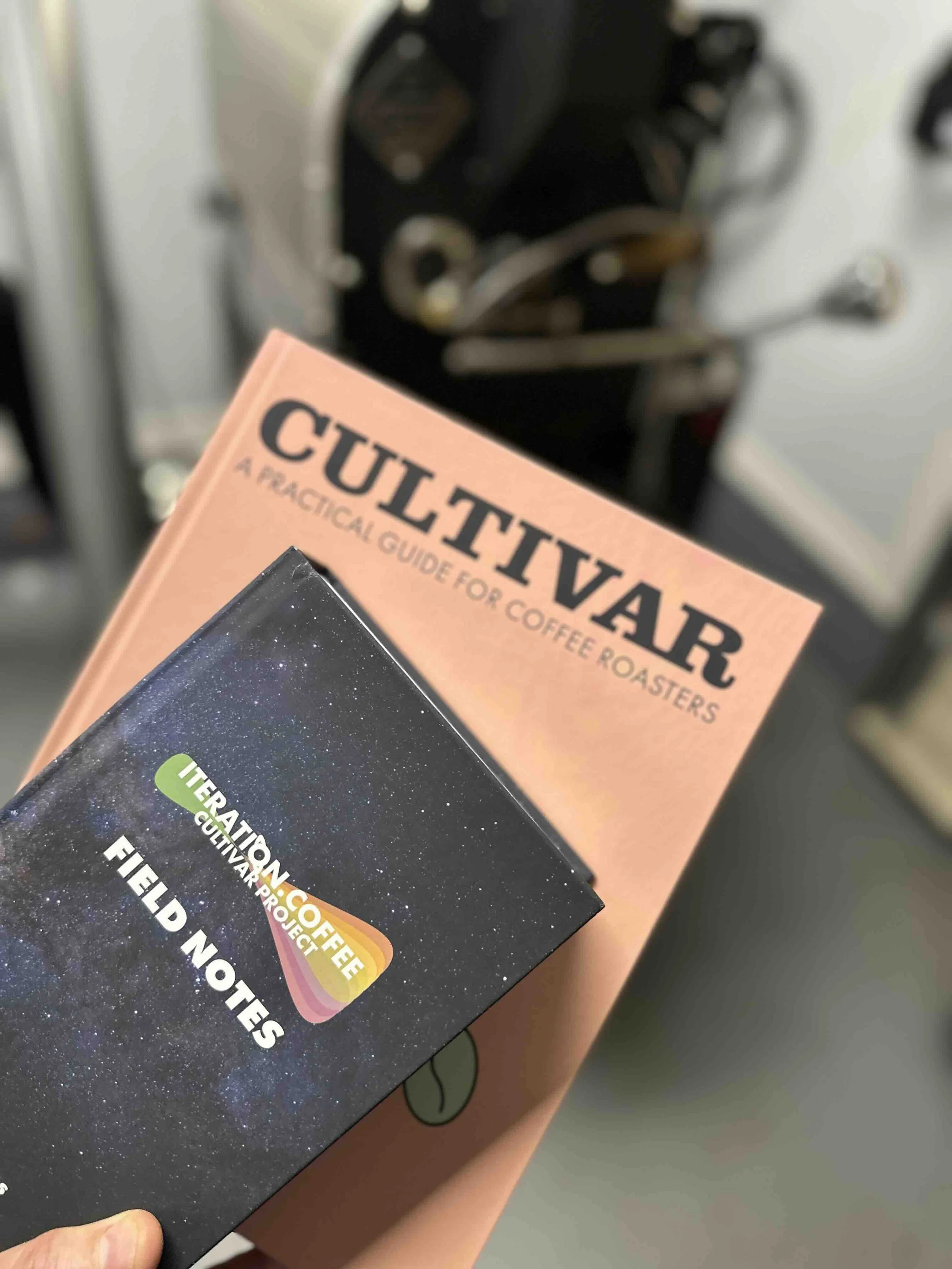 A person holding a book titled 'CULTIVAR' and a black notebook with the logo 'INTERATION COFFEE CULTIVAR PROJECT FIELD NOTES' in front of a coffee roasting machine.