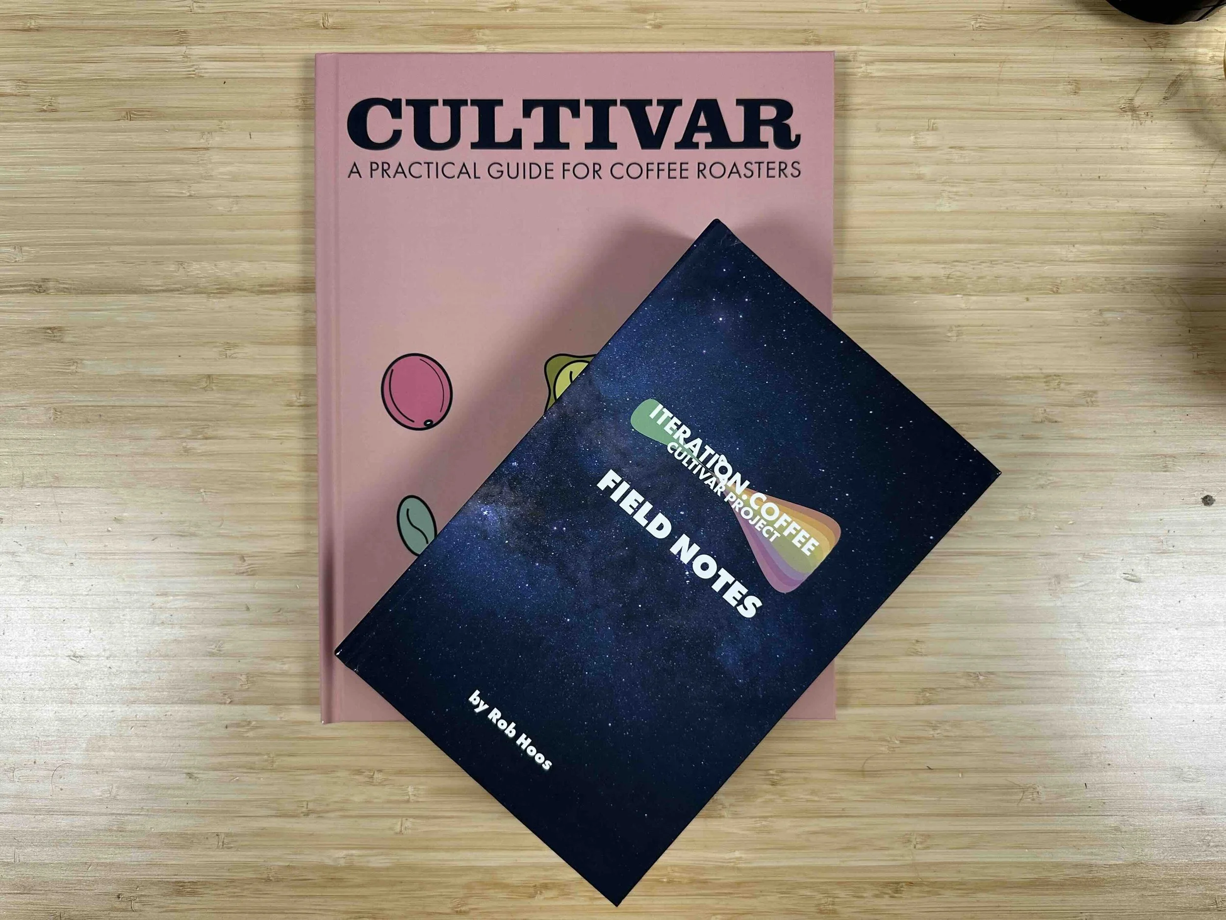 A pink book titled "Cultivar: A Practical Guide for Coffee Roasters" and a dark blue notebook titled "Field Notes" by Rob Hoos on a wooden surface.