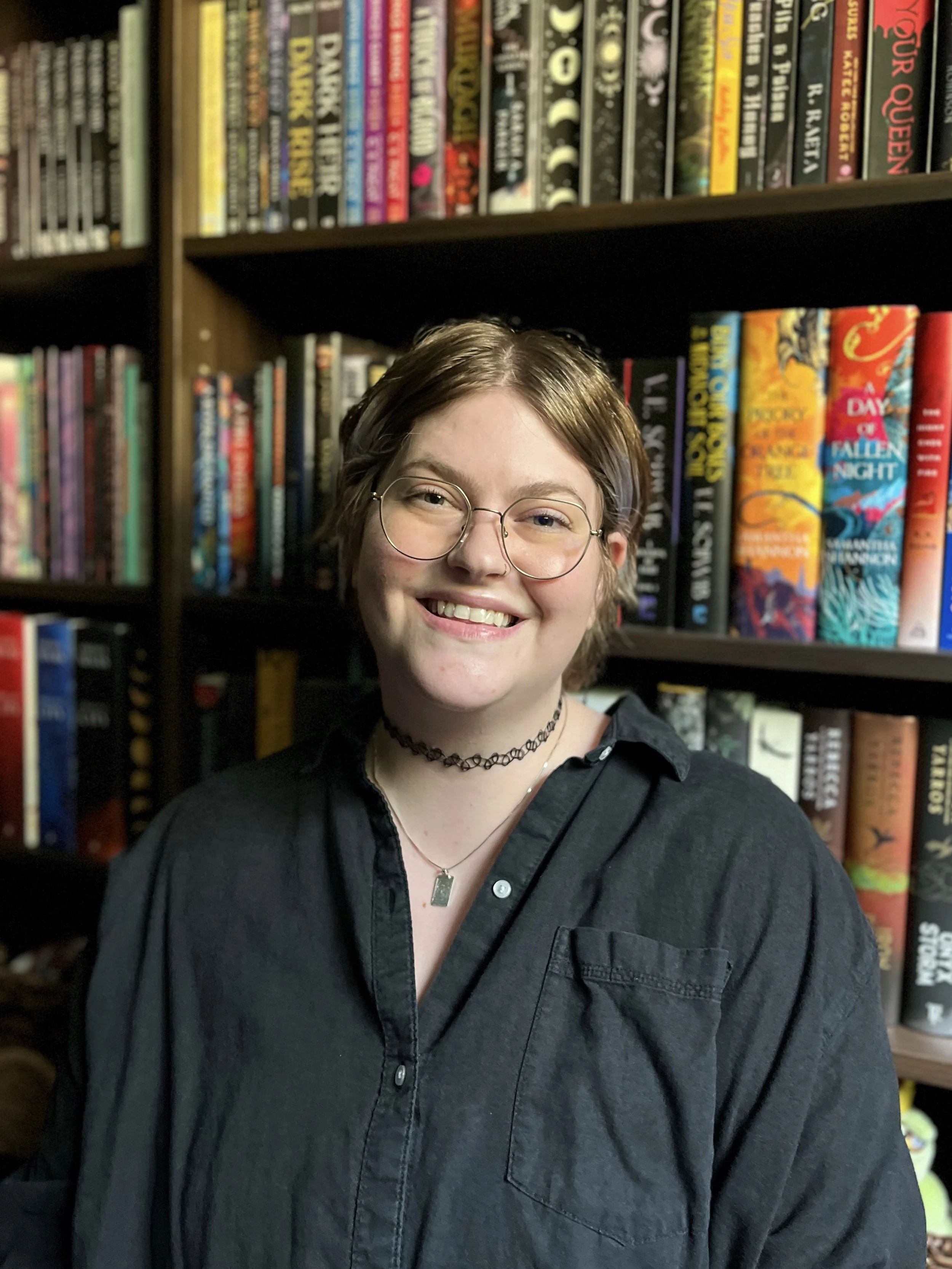 A person with short, blue and gray hair wearing glasses, a black shirt, and necklaces, smiling in front of a bookshelf filled with colorful books.