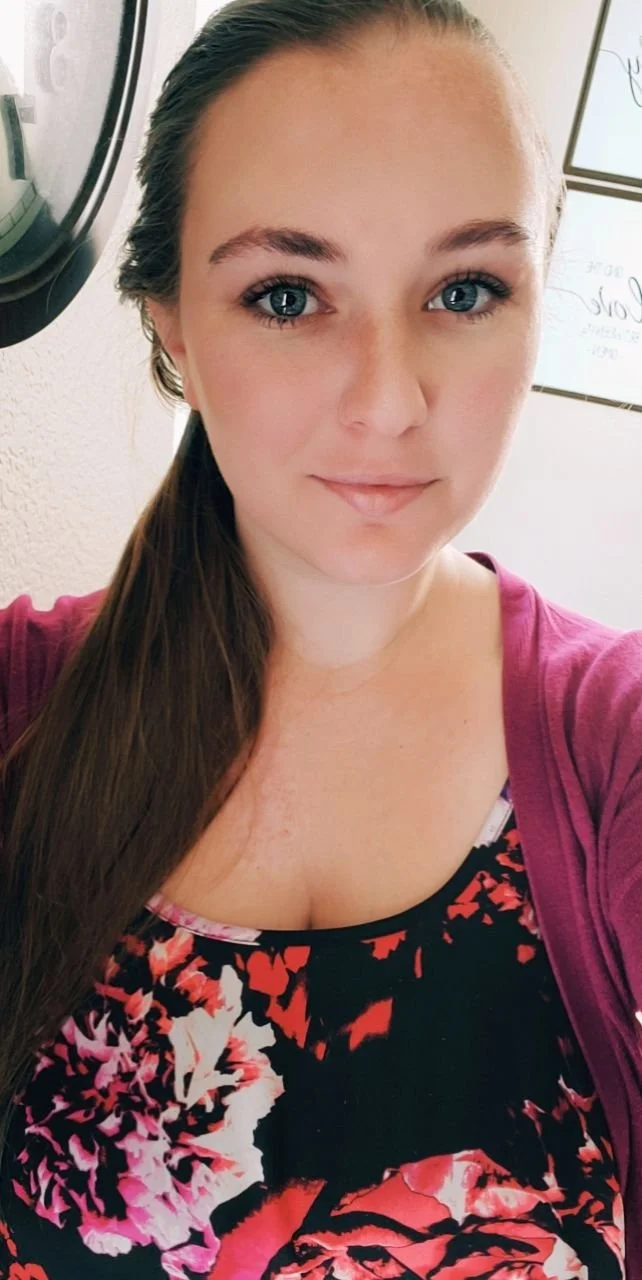 Close-up portrait of a young woman with fair skin, blue eyes, and long brown hair, wearing a floral top and maroon cardigan, standing indoors with  artwork in the background.