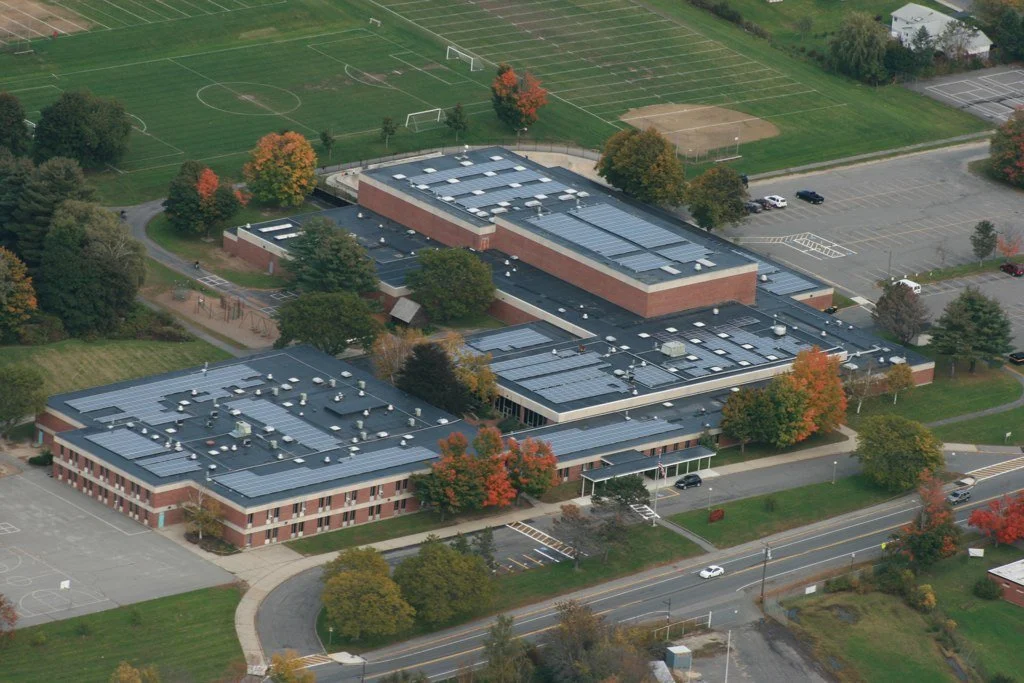 Aerial view of a school building complex with solar panels on the roof, sports fields, parking lots, and surrounding trees with fall foliage.