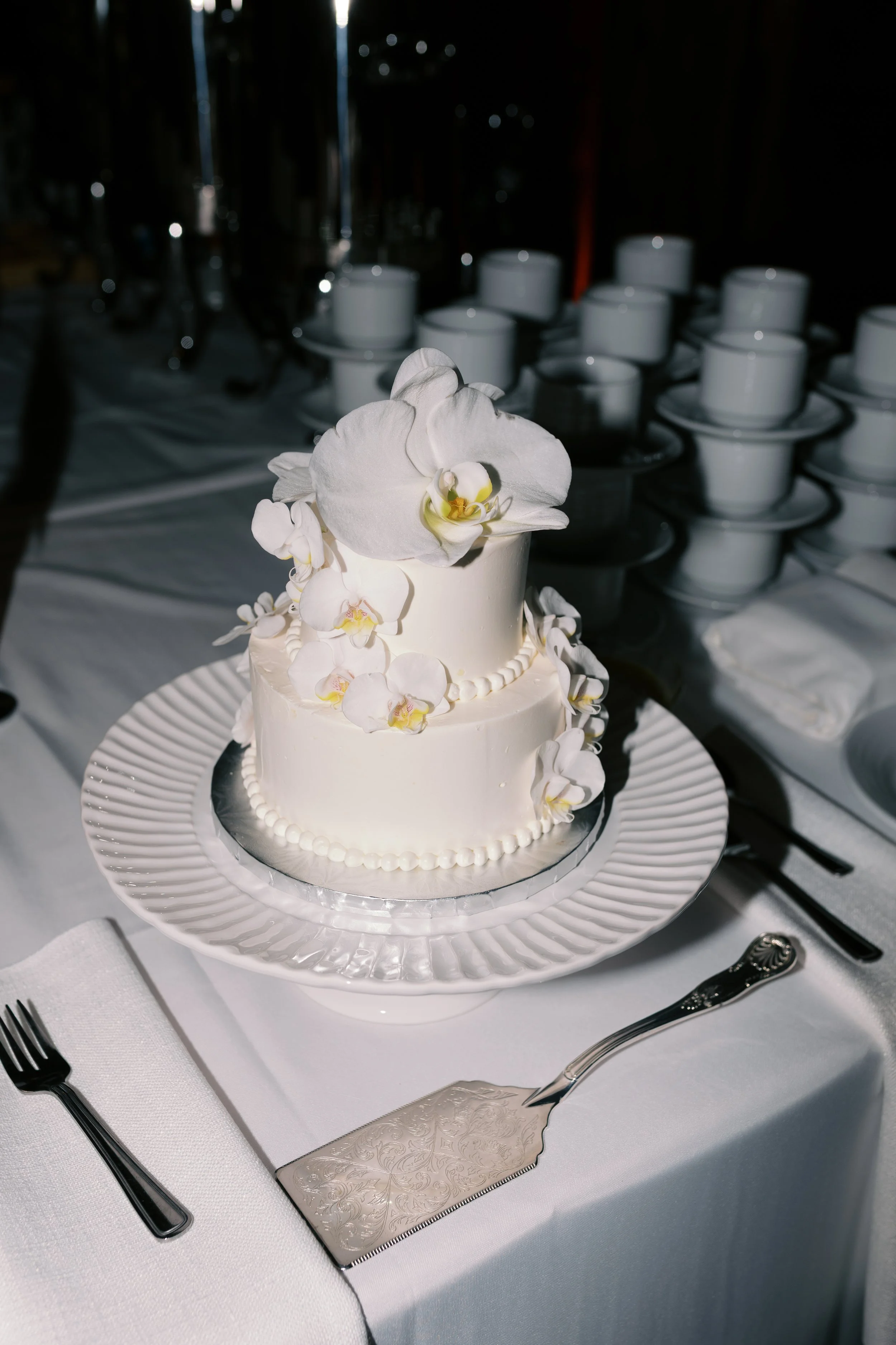 Two-tier white wedding cake decorated with white orchids, placed on a white tablecloth at a formal event.
