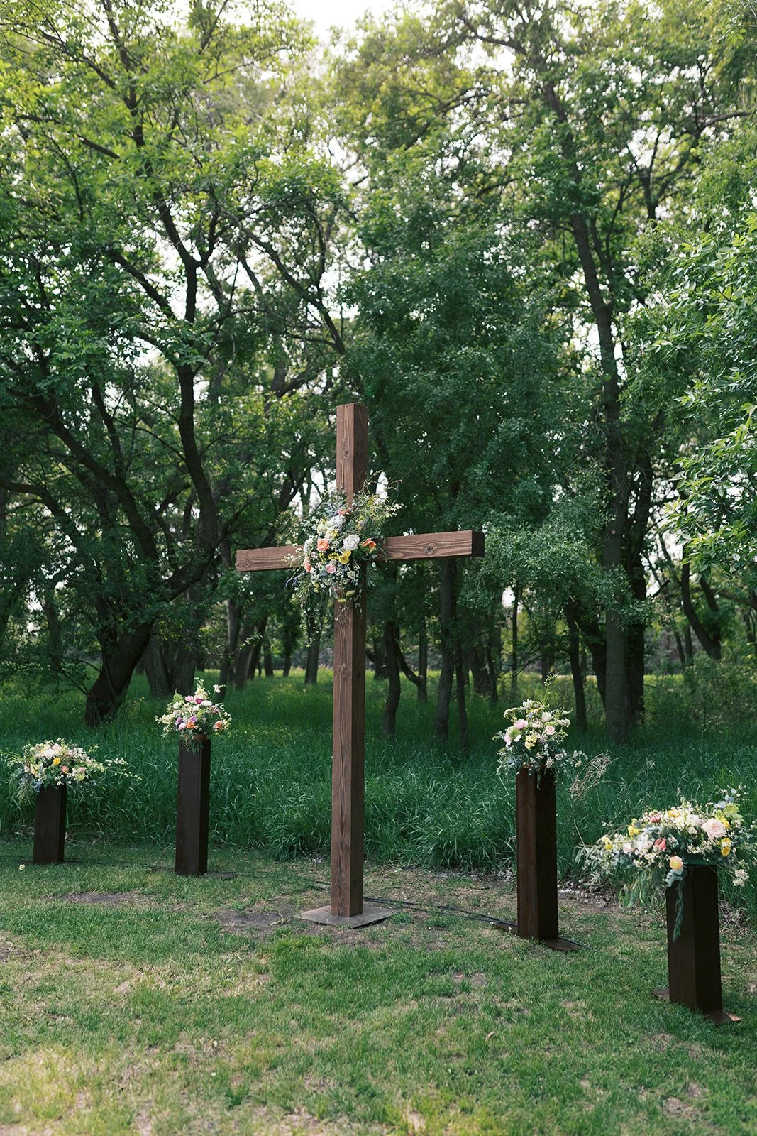 An outdoor wedding setup with a wooden cross adorned with flowers, surrounded by four flower arrangements on dark pedestals, set in a green, wooded area.