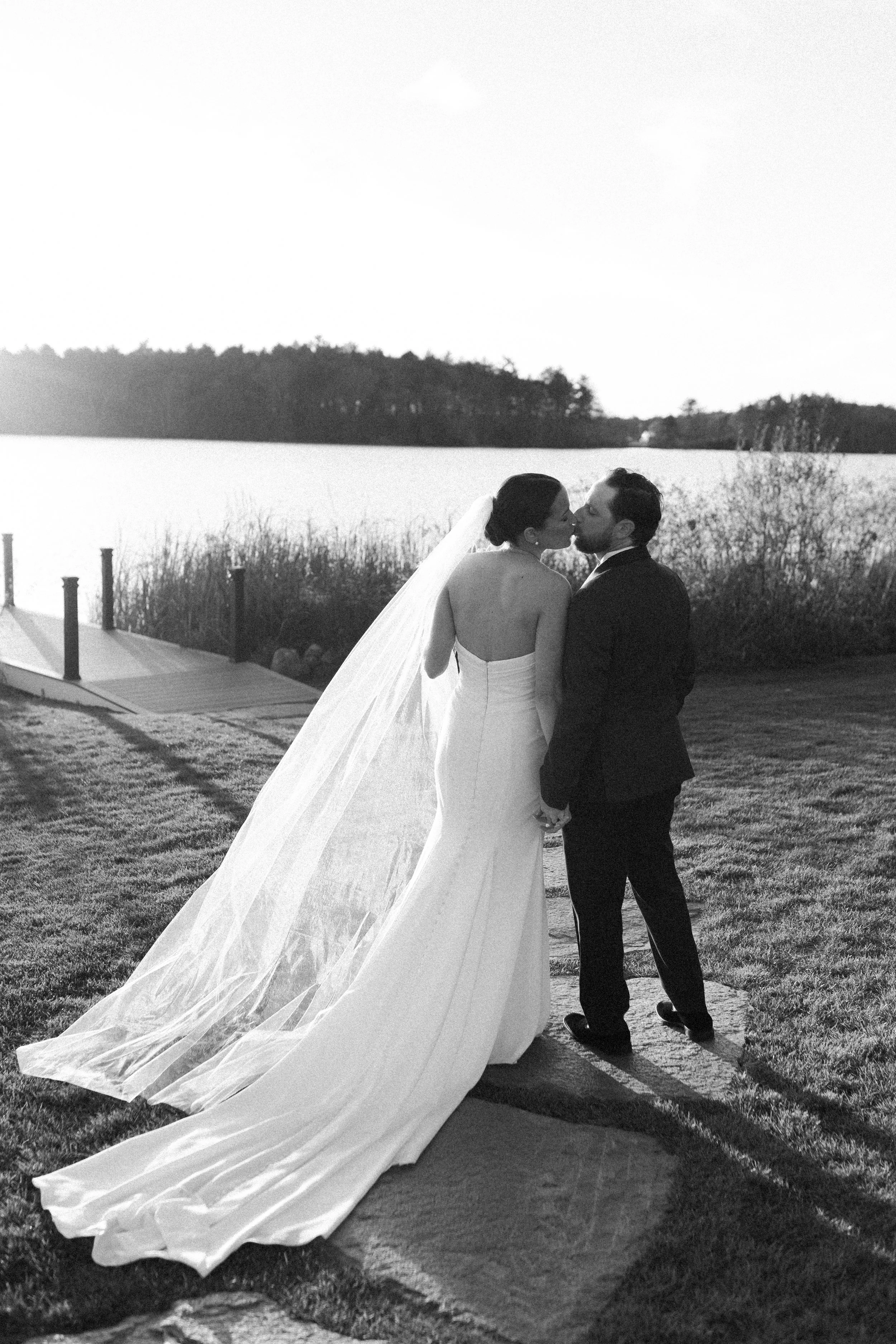 A bride and groom kissing outdoors by a lake at sunset, with the bride in a strapless gown with a long train and veil, and the groom in a suit.