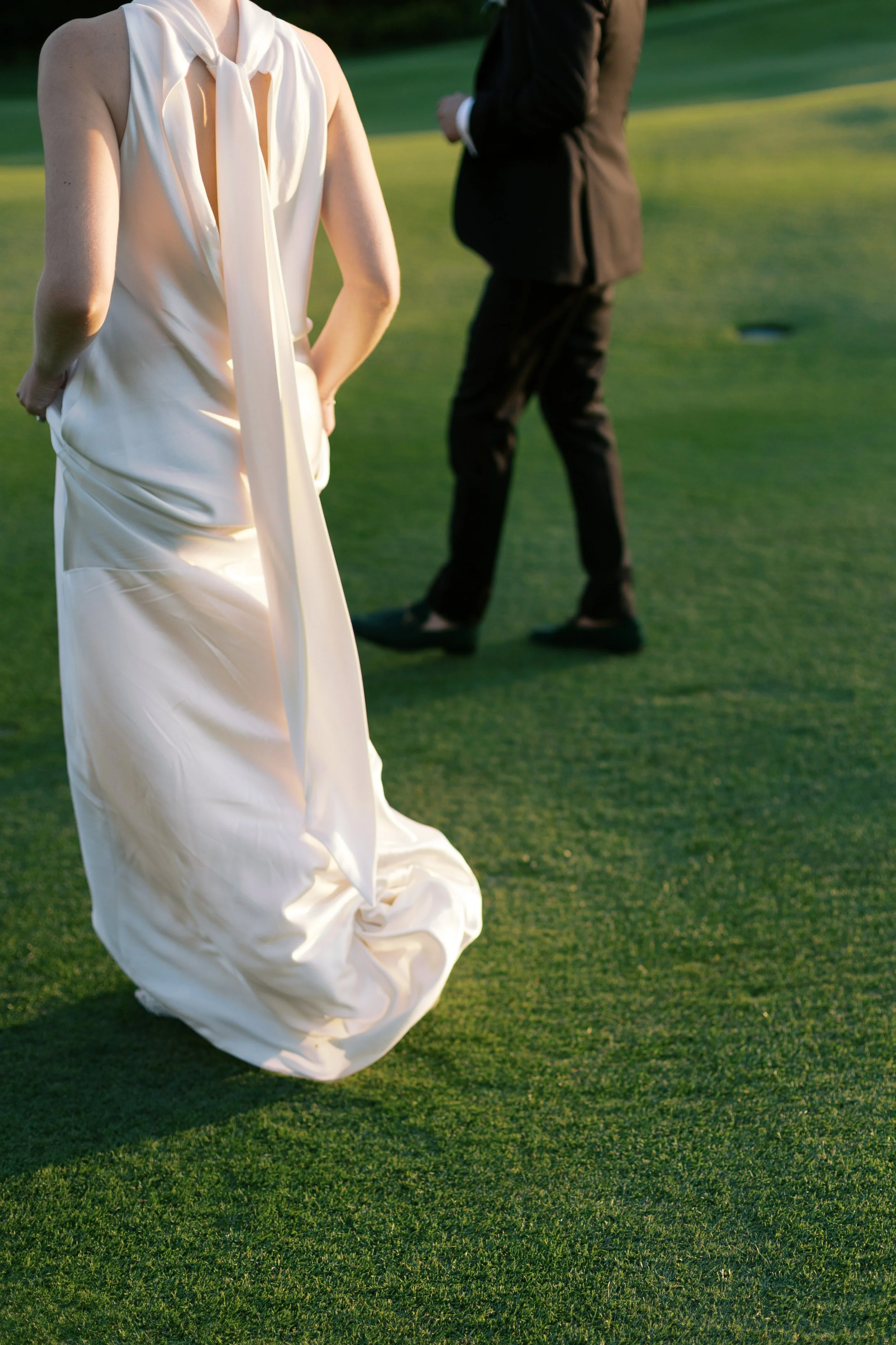 A woman in a white dress stands on a golf course with her hands behind her back, and a man in a black suit stands in the background on the green grass.