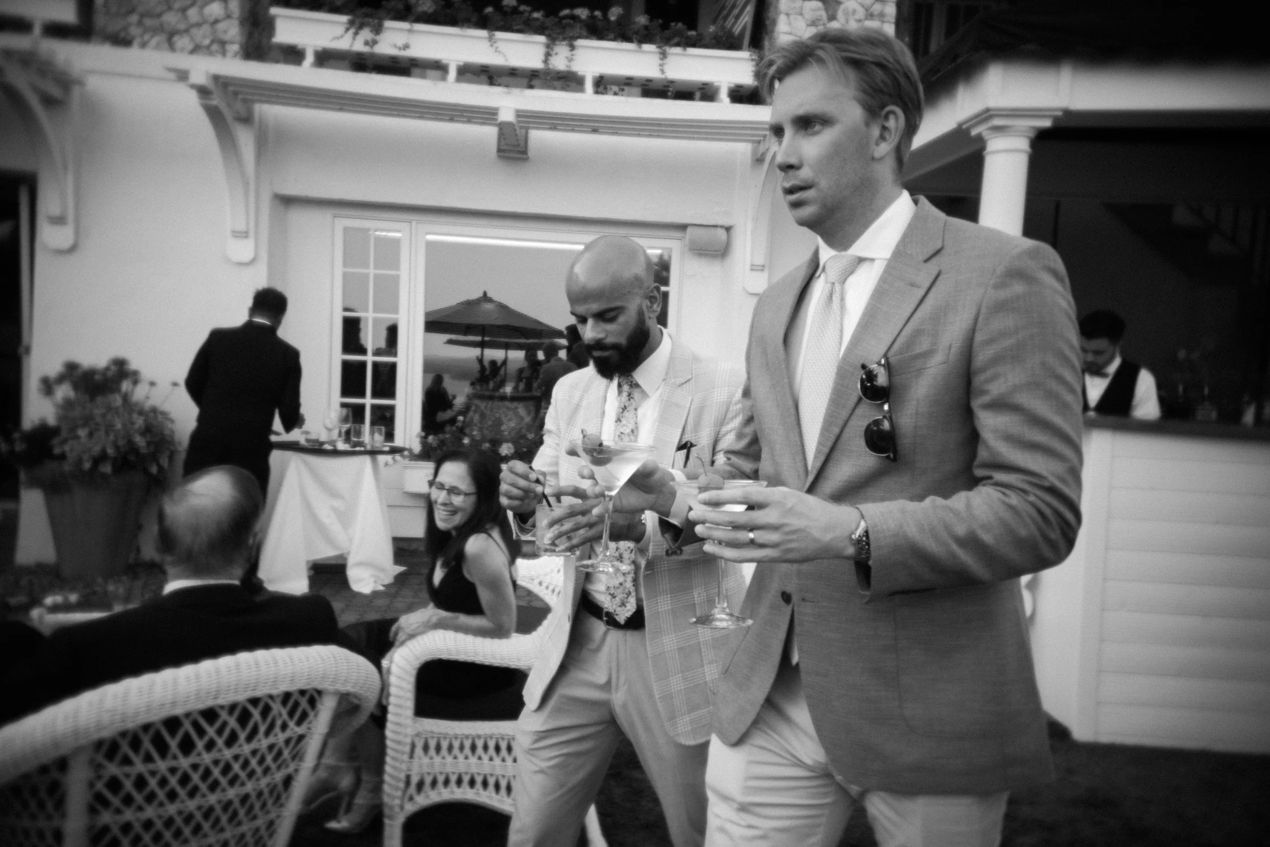 People at a social gathering, two men wearing suits holding cocktails, woman sitting at a table smiling, outdoor setting with a bar and decorative plants in the background.
