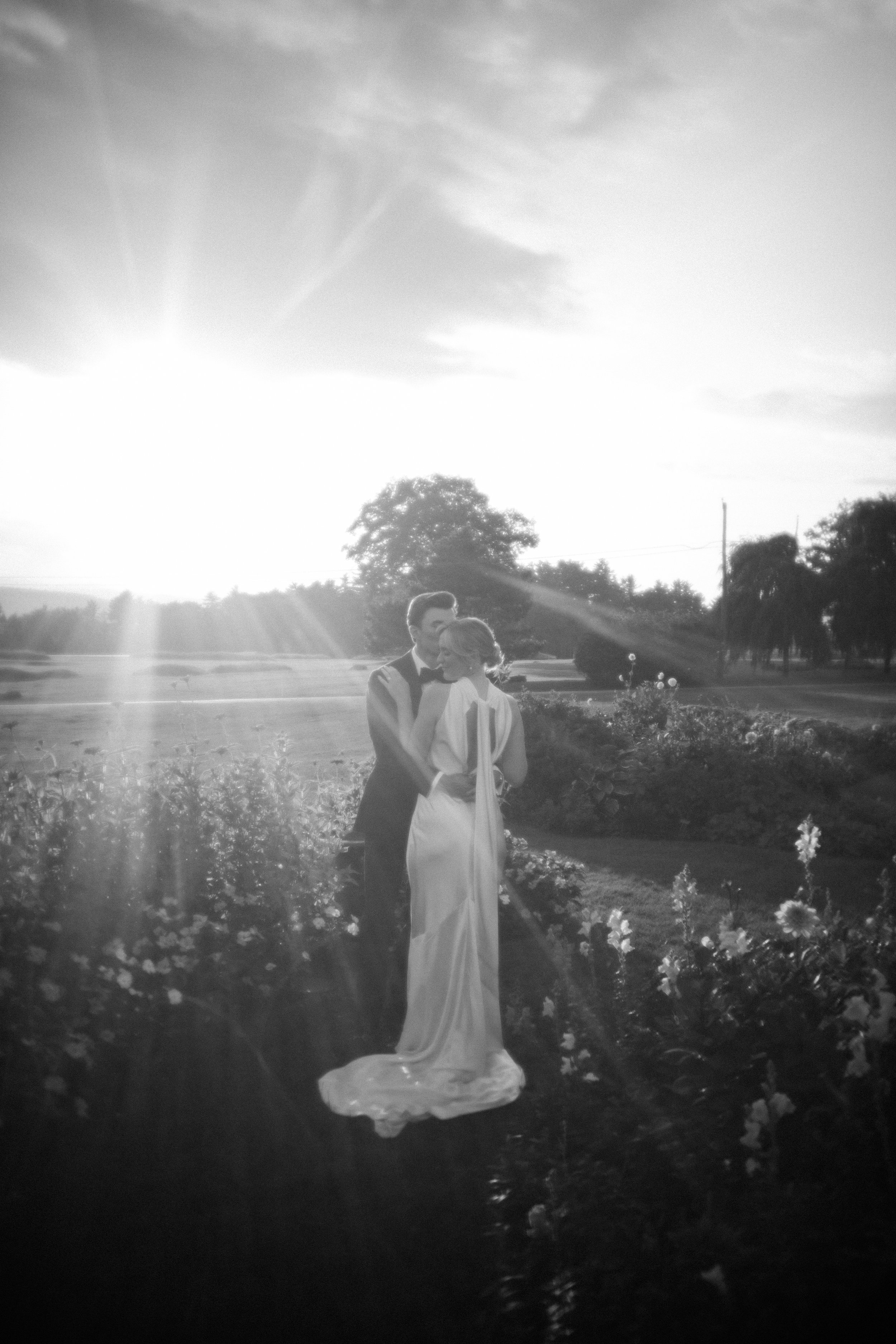 A black-and-white photo of a couple in wedding attire embracing in a garden with sunlight shining through the trees.