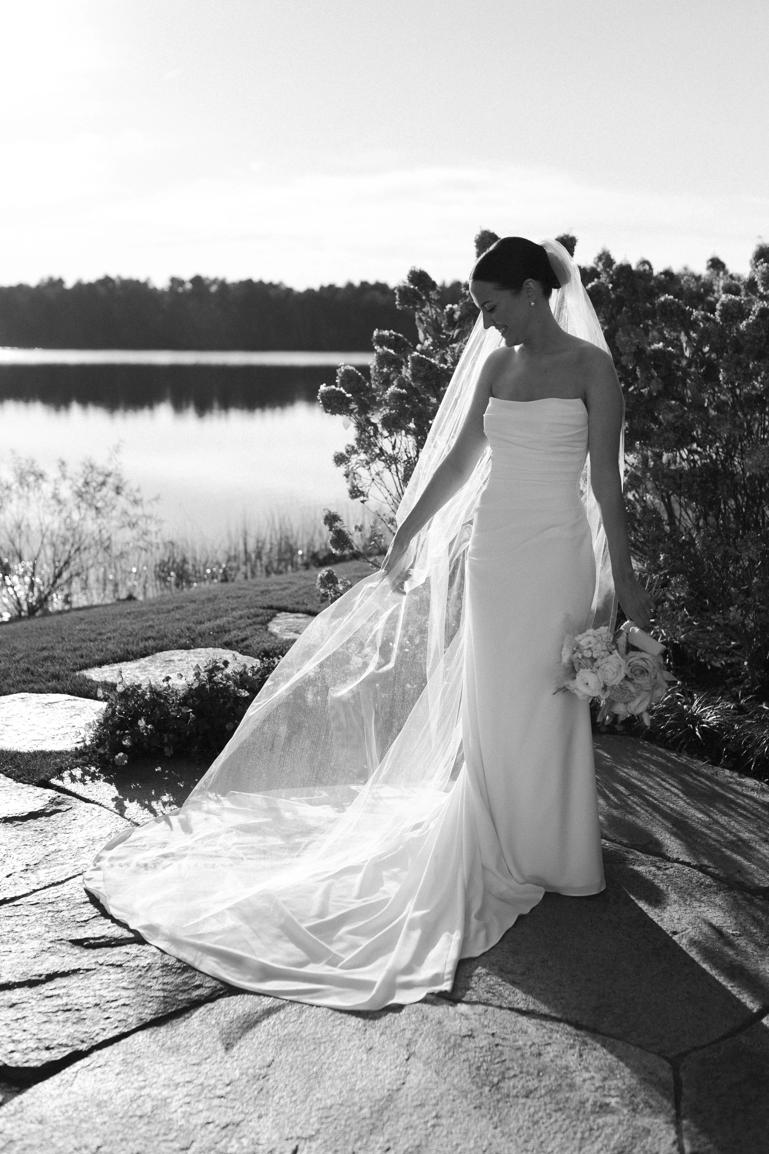 A bride in a strapless wedding dress holding a bouquet of flowers, standing on a stone path outdoors near a lake, with bushes and trees in the background.