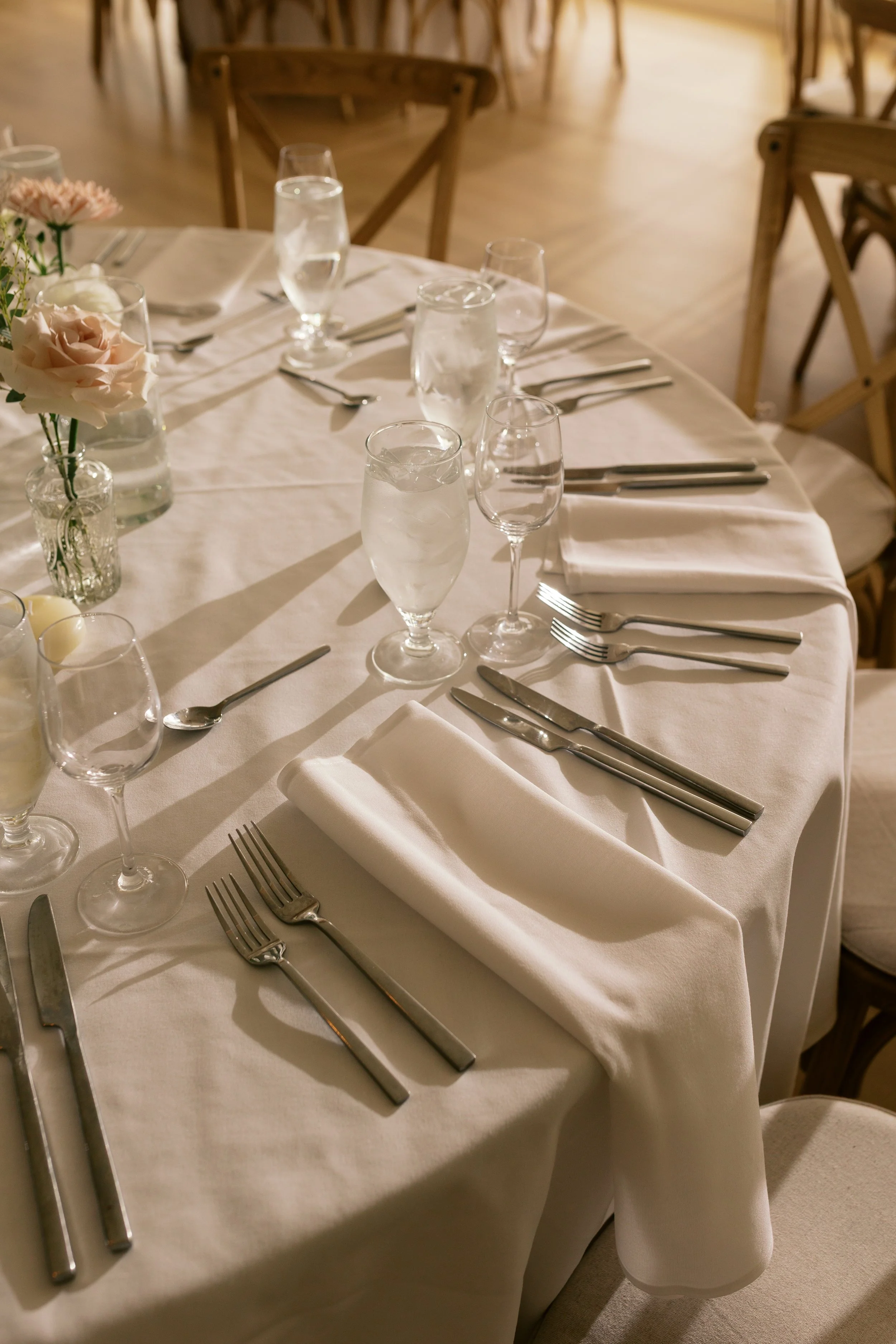 A round banquet table set for a formal event with white tablecloth, neatly folded white napkins, multiple wine glasses, water glasses with ice, and silverware arranged for a meal. The table is decorated with pink and white flowers in glass vases.