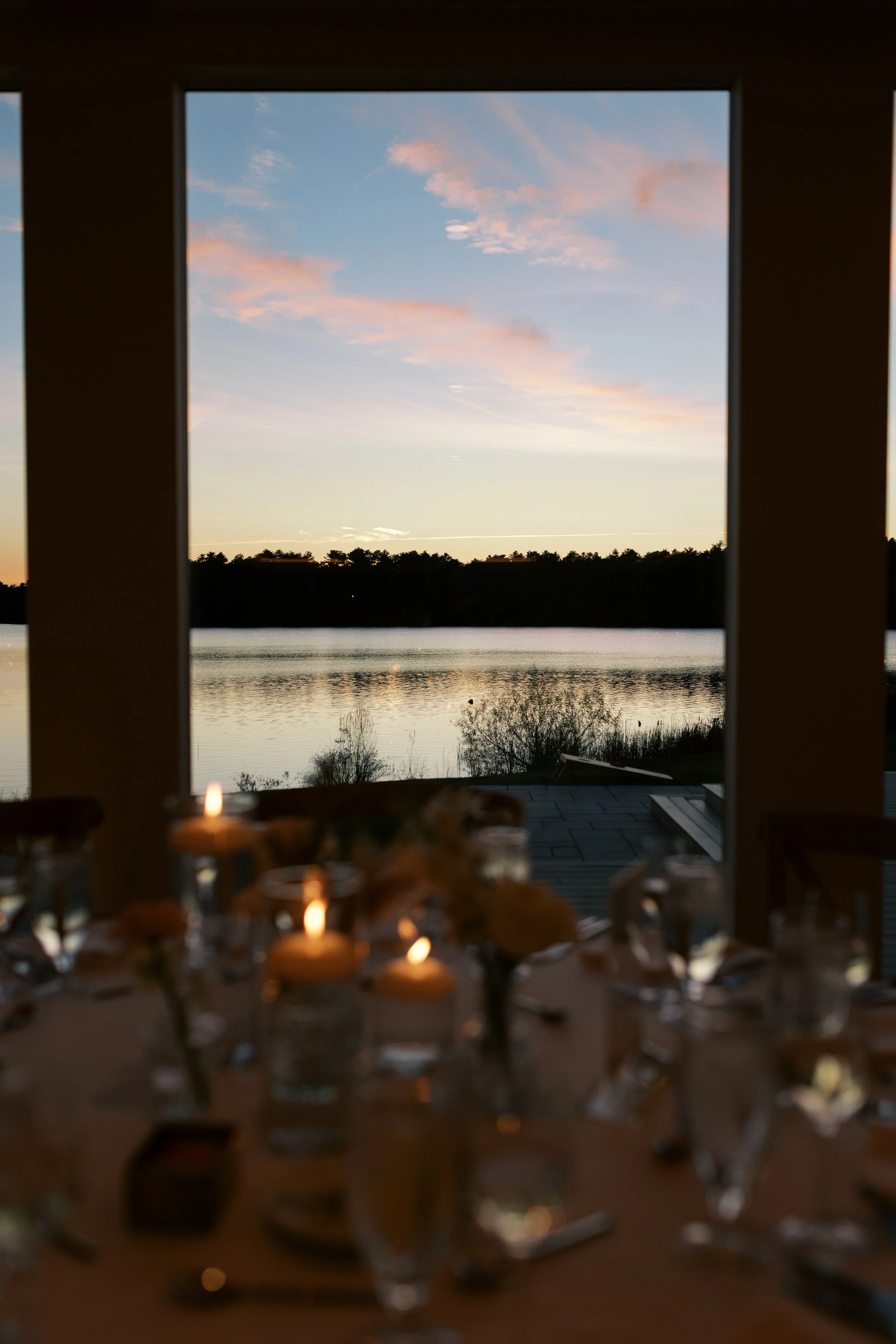 A lakeside view through a window at sunset with a decorated table, softly lit with candles, in the foreground.