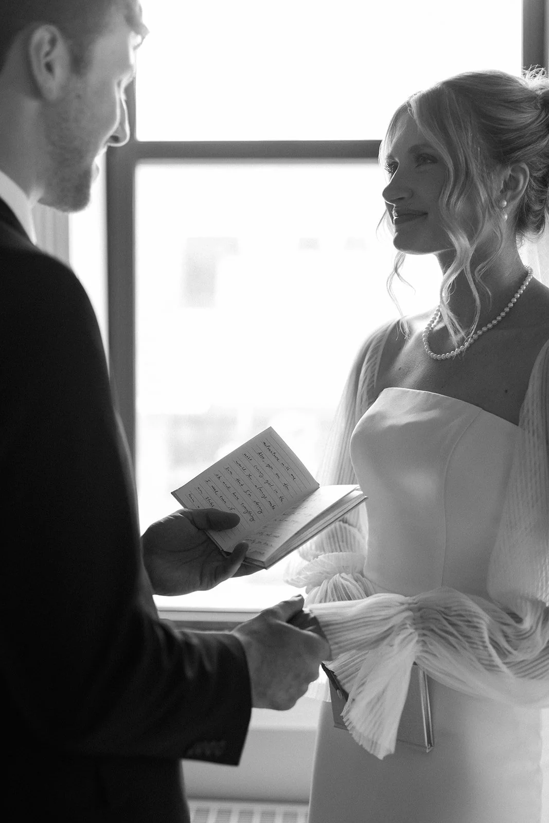A man and woman standing by a window during a wedding ceremony, with the man holding a small book or notepad, and the woman smiling at him. The woman is dressed in a wedding gown with a pearl necklace, and the man in a suit.