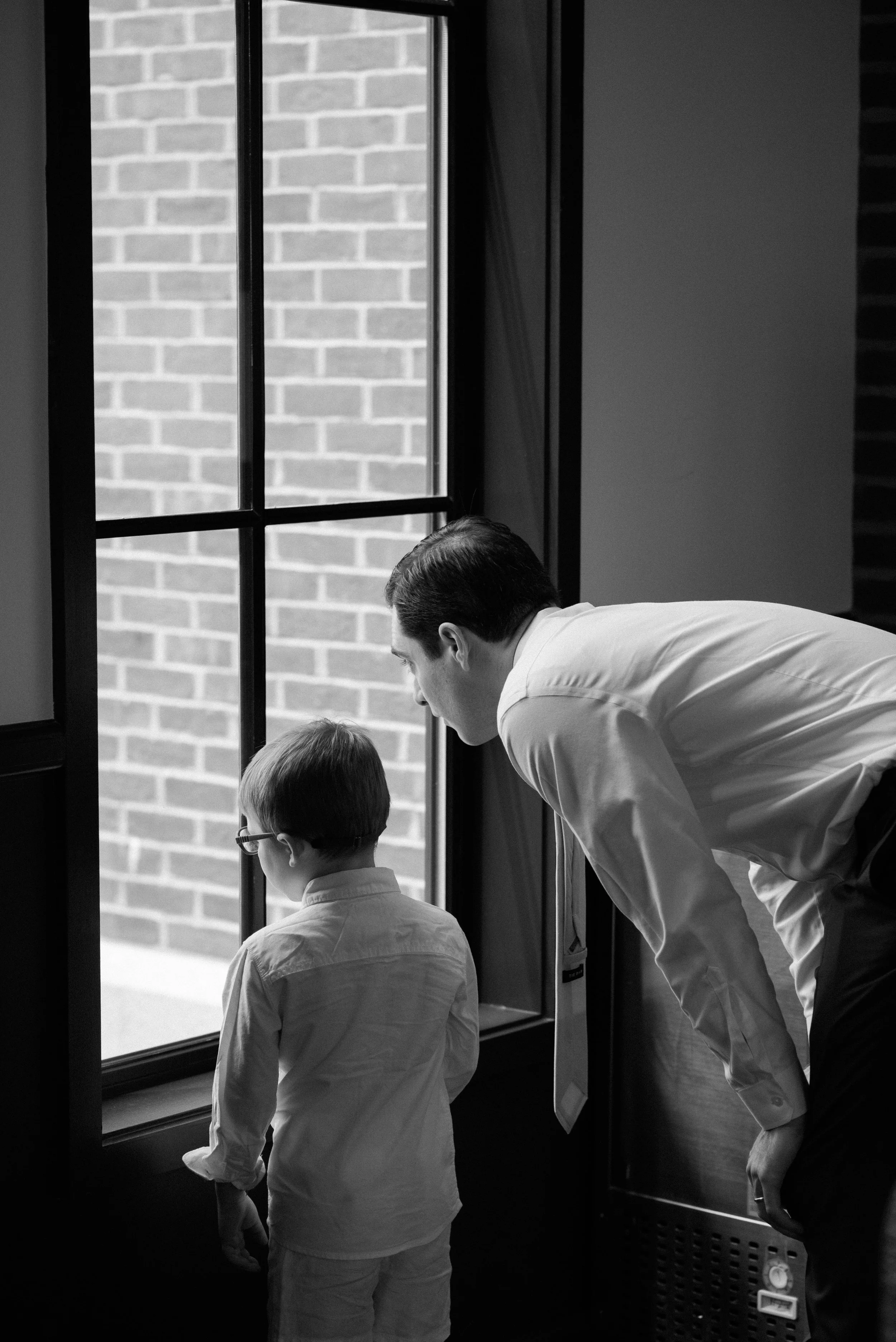 A man and a young boy looking out of a large window with brick exterior wall visible outside, both dressed in formal attire.