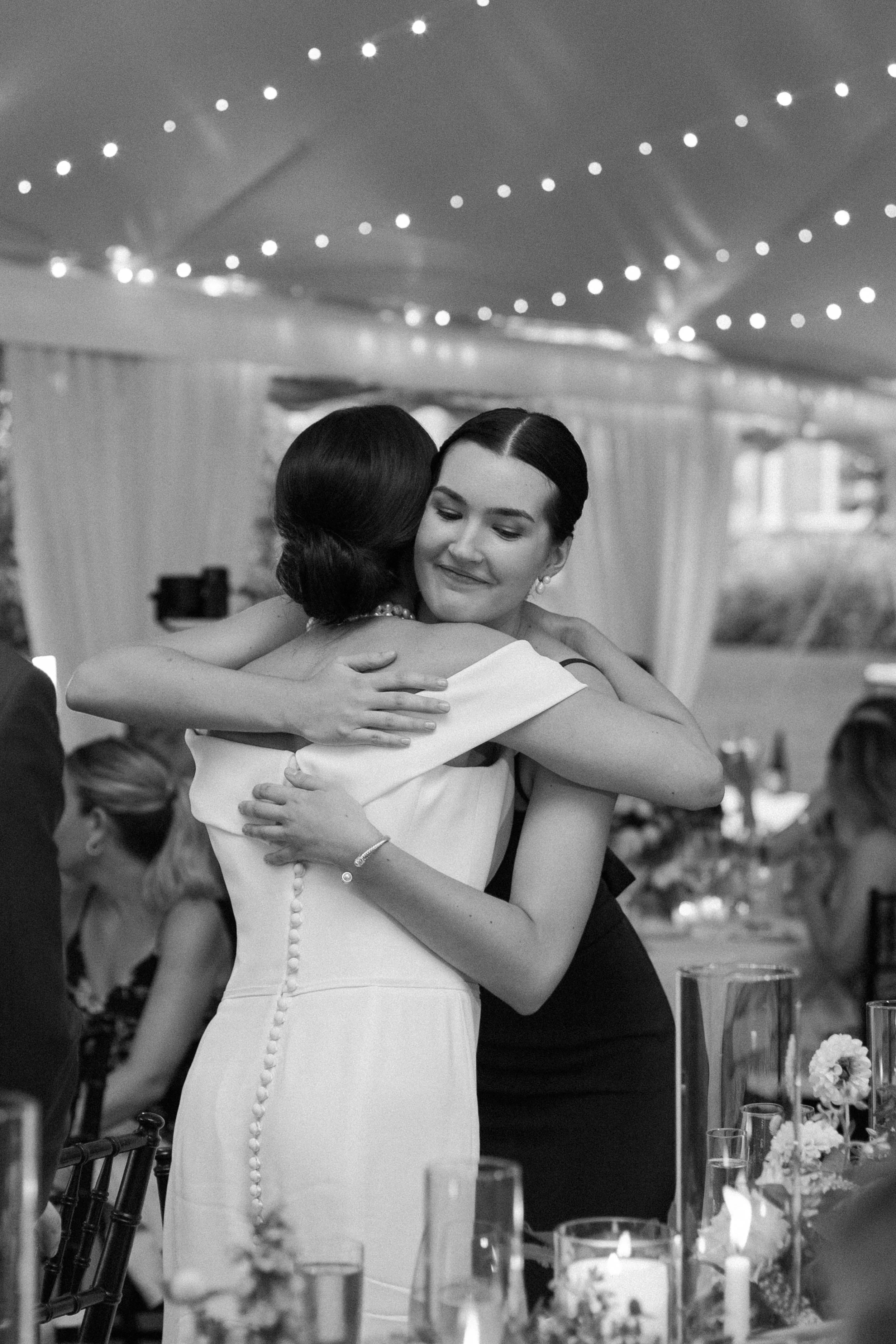 Two women hugging at a wedding reception, one in a white dress and the other in a dark dress, under string lights.