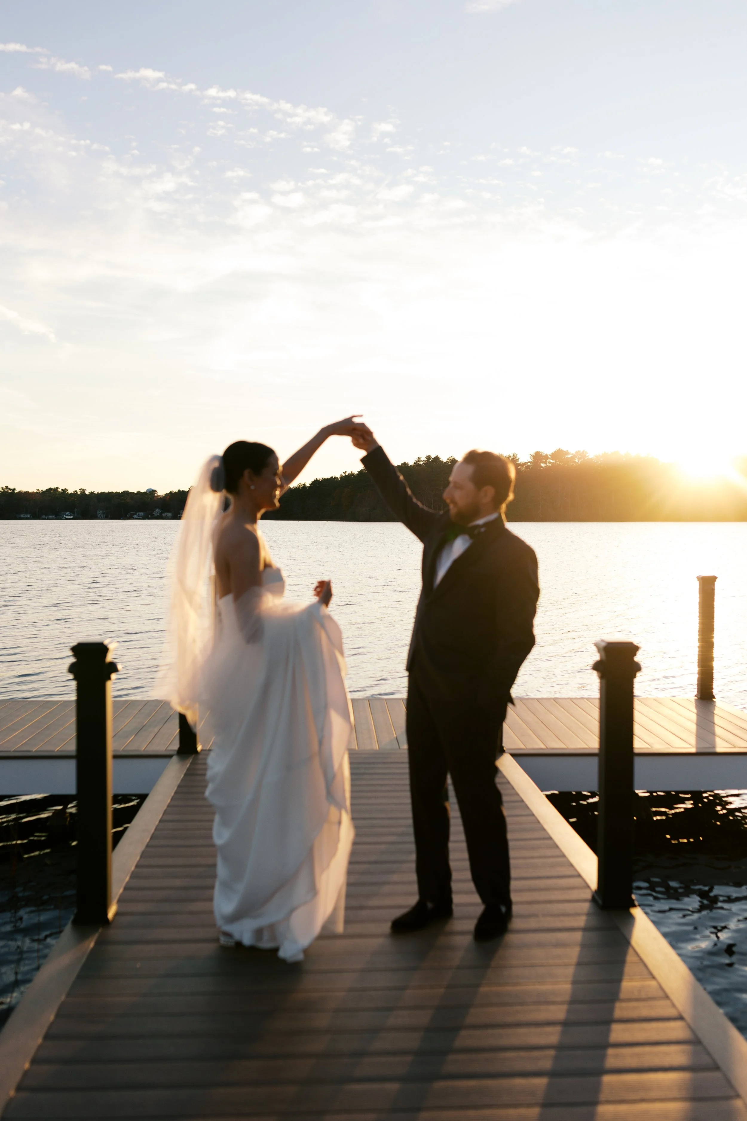 A bride and groom dancing on a dock by a lake during sunset, with the groom twirling the bride.