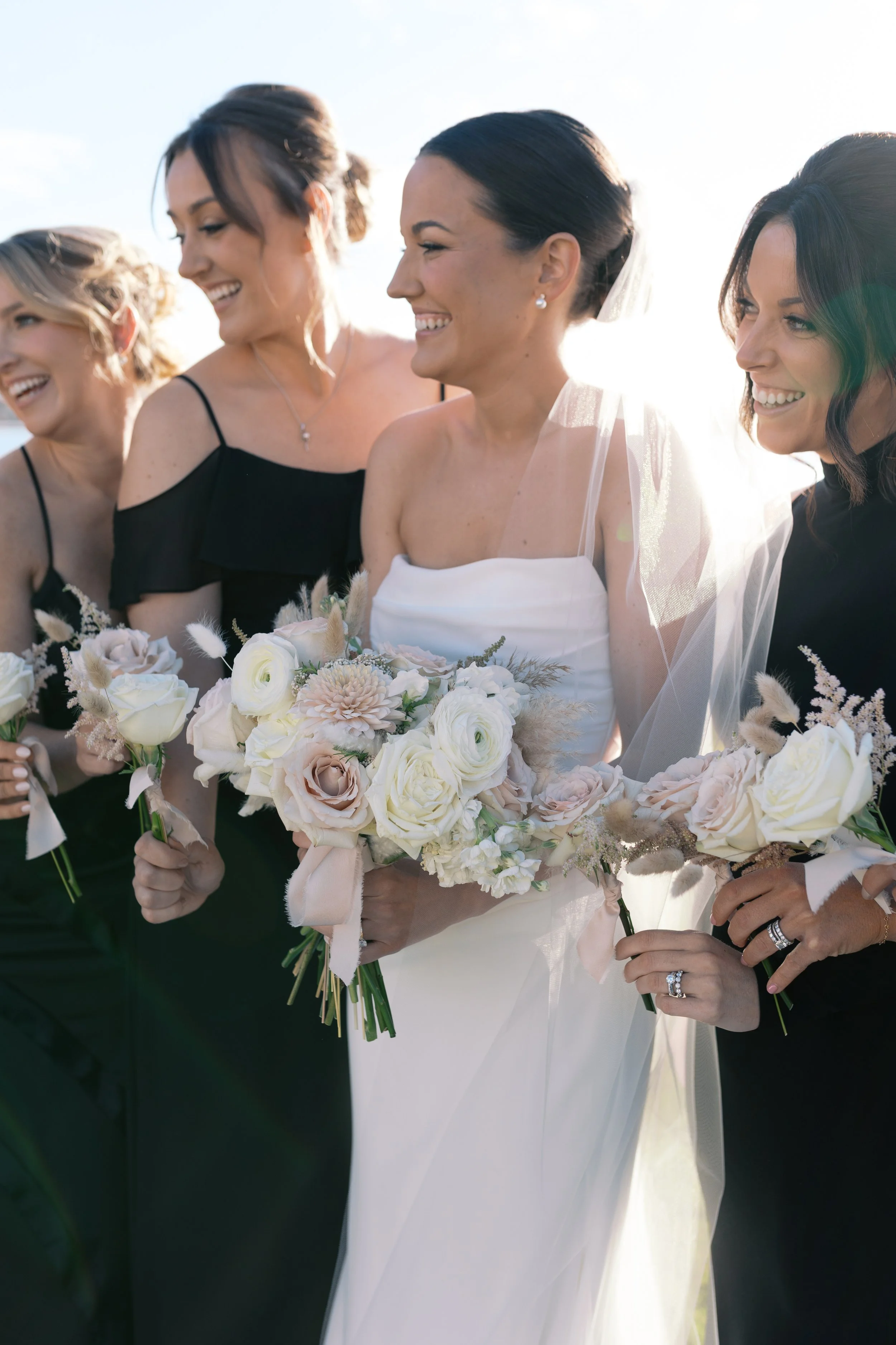 Bride in white dress holding a bouquet of white and blush flowers, surrounded by four women in black dresses, all smiling outdoors with sunlight.
