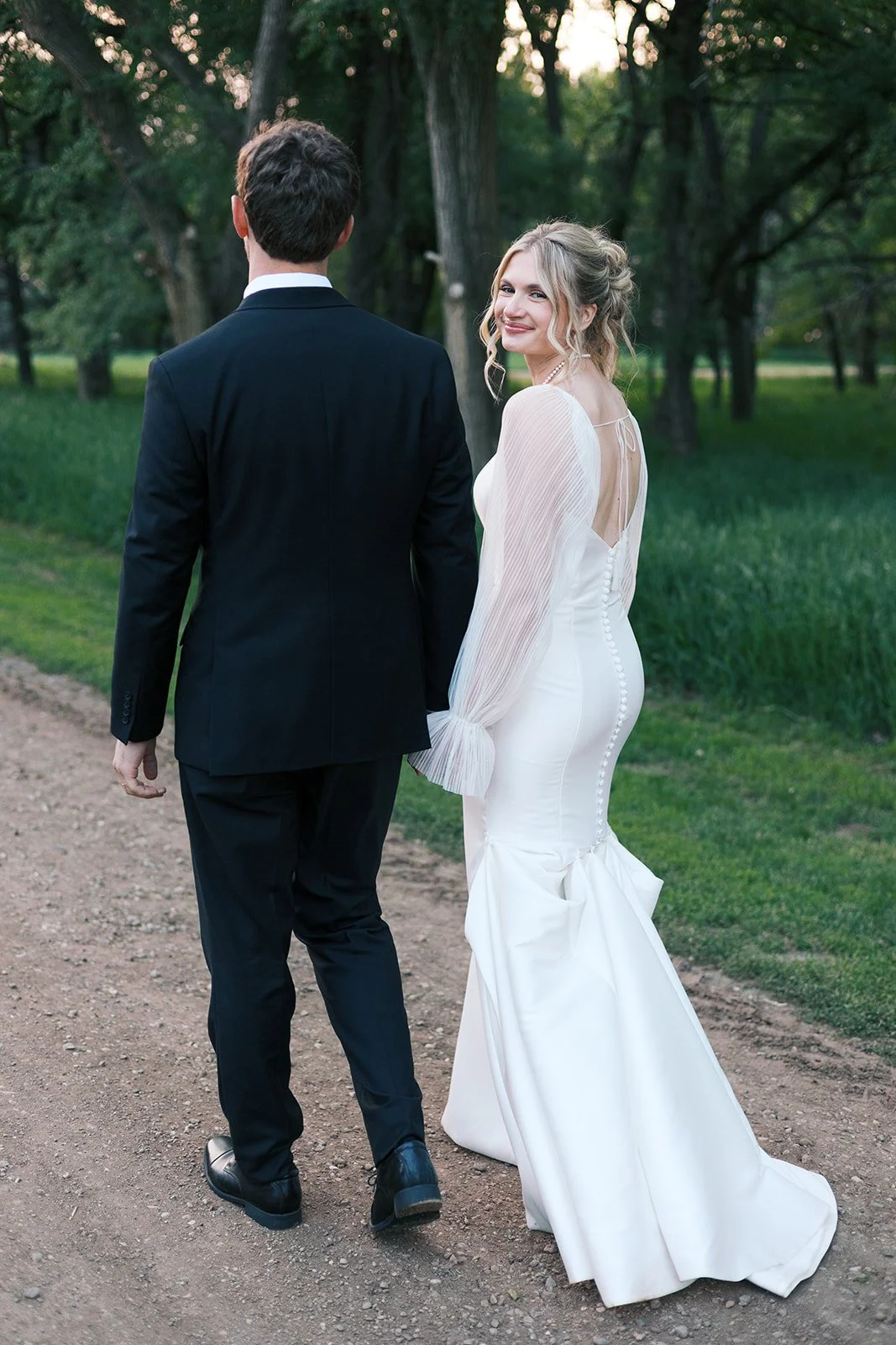A couple dressed in formal wedding attire, walking hand in hand outdoors on a dirt path surrounded by greenery and trees, with the bride smiling at the camera.