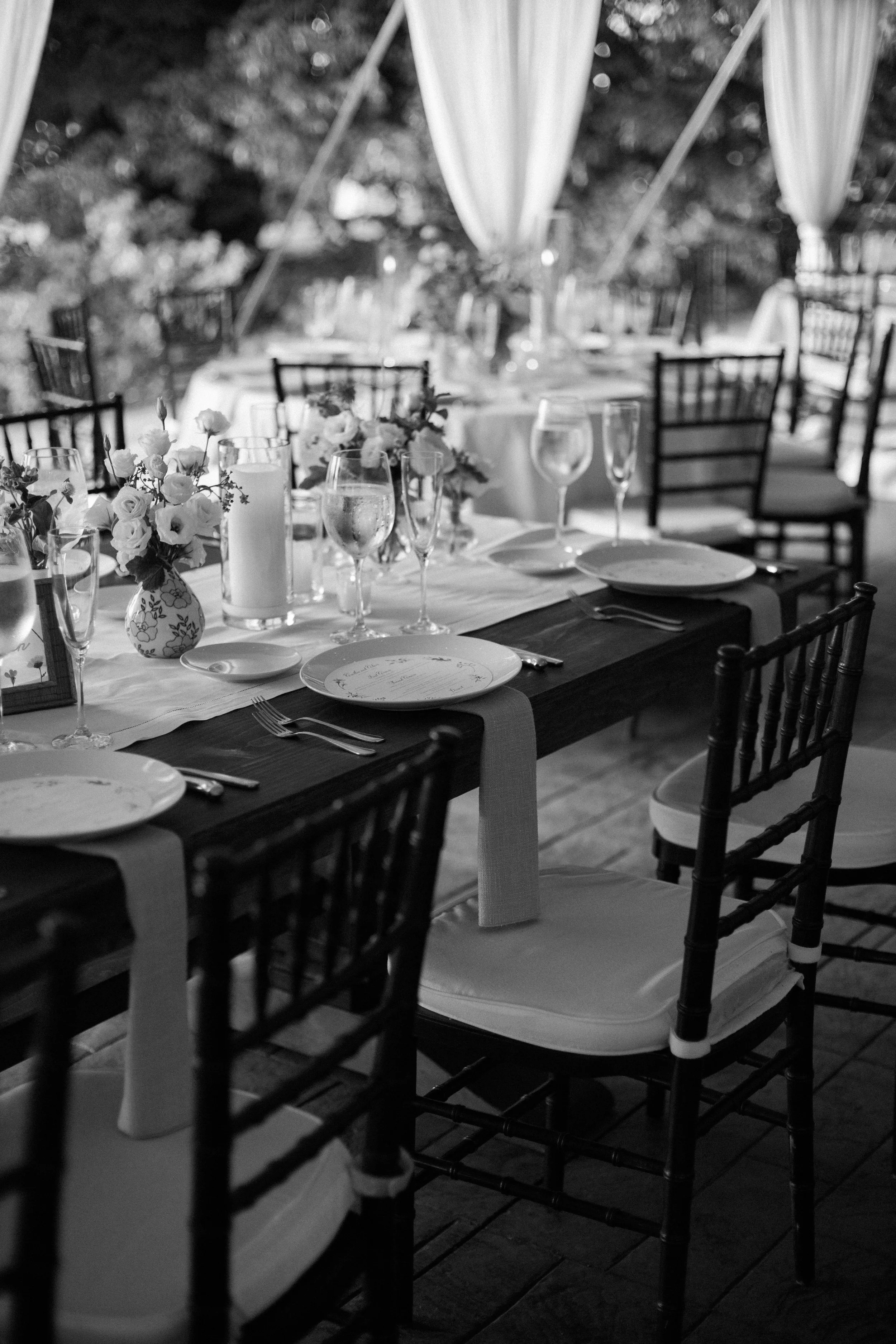 Elegant outdoor dining table set for a celebration, decorated with flowers, glassware, plates, and candles, with draped curtains and lush greenery in the background.