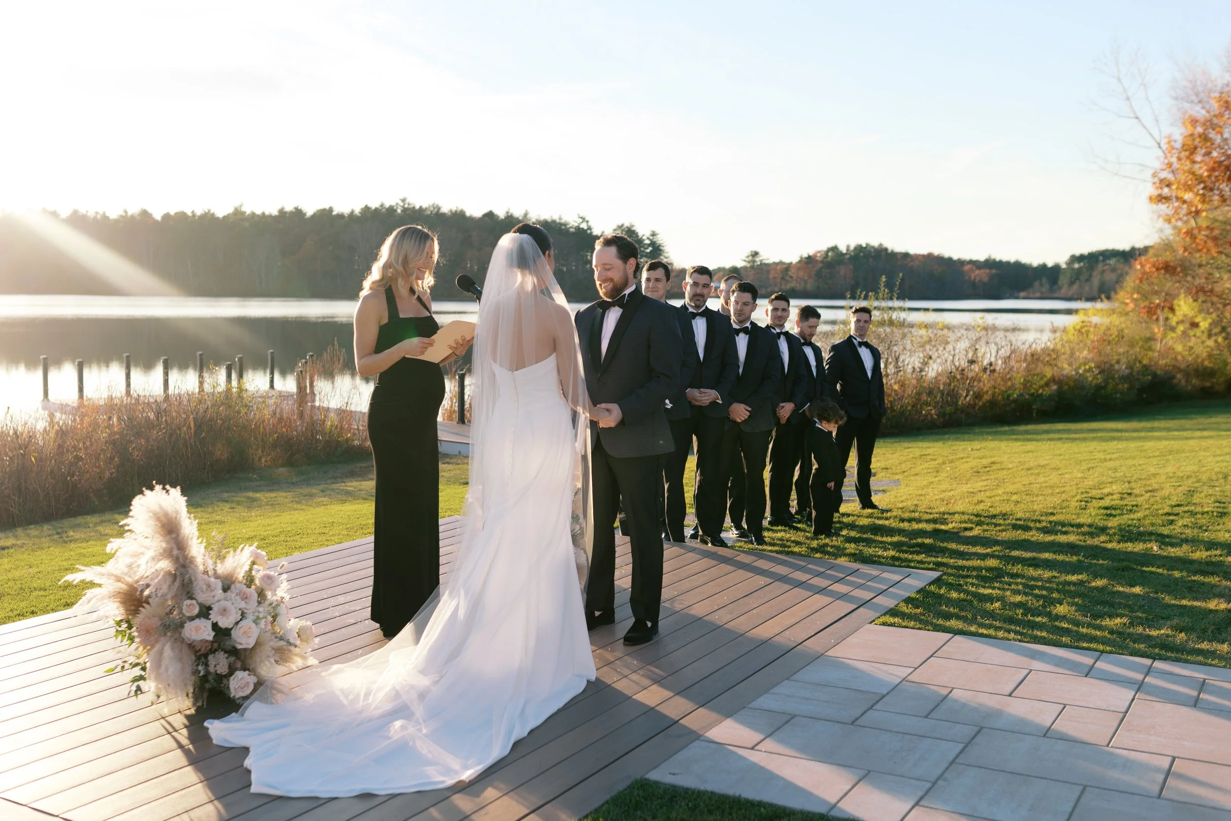 A wedding ceremony taking place outdoors by a lake with a officiant reading from a paper, a bride and groom holding hands, and groomsmen standing in line on the grass, during sunset.