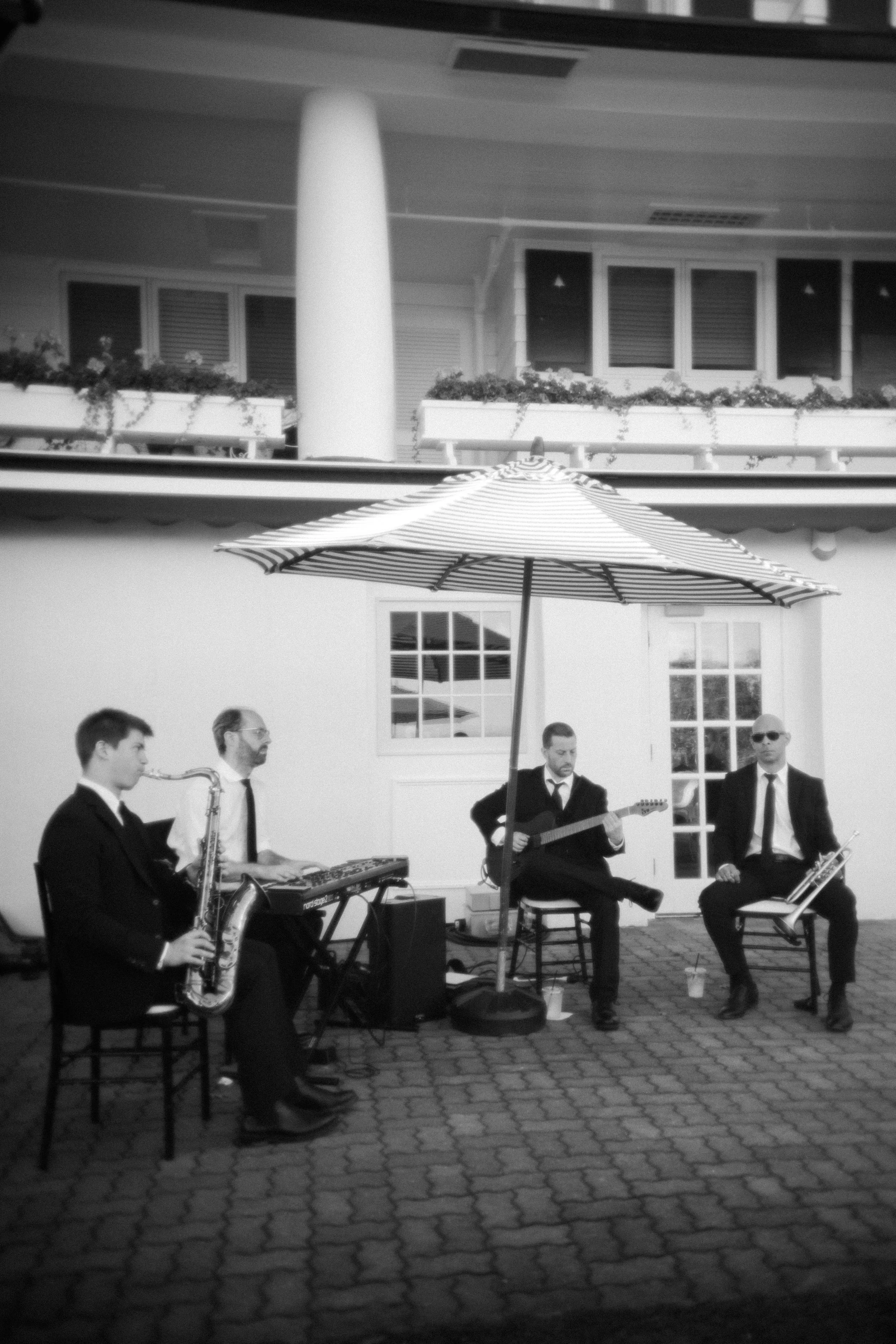Four musicians in suits perform outdoors under a large striped umbrella, with one playing the keyboard, another playing the guitar, a third playing the saxophone, and the fourth playing the trumpet, in front of a building with windows and balcony.