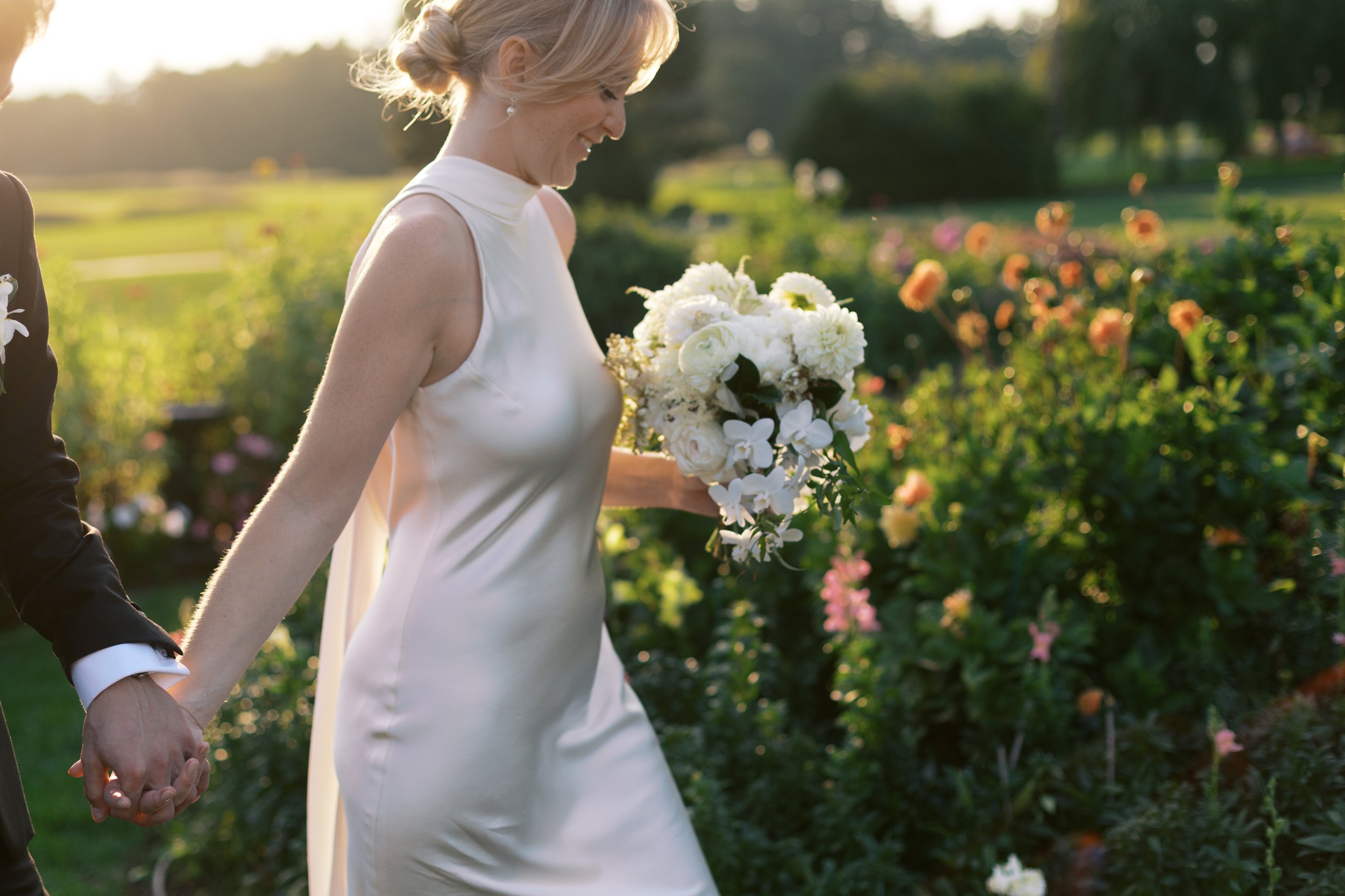 A bride in a white wedding dress holding a bouquet of white flowers, walking hand-in-hand with her partner in a garden at sunset.