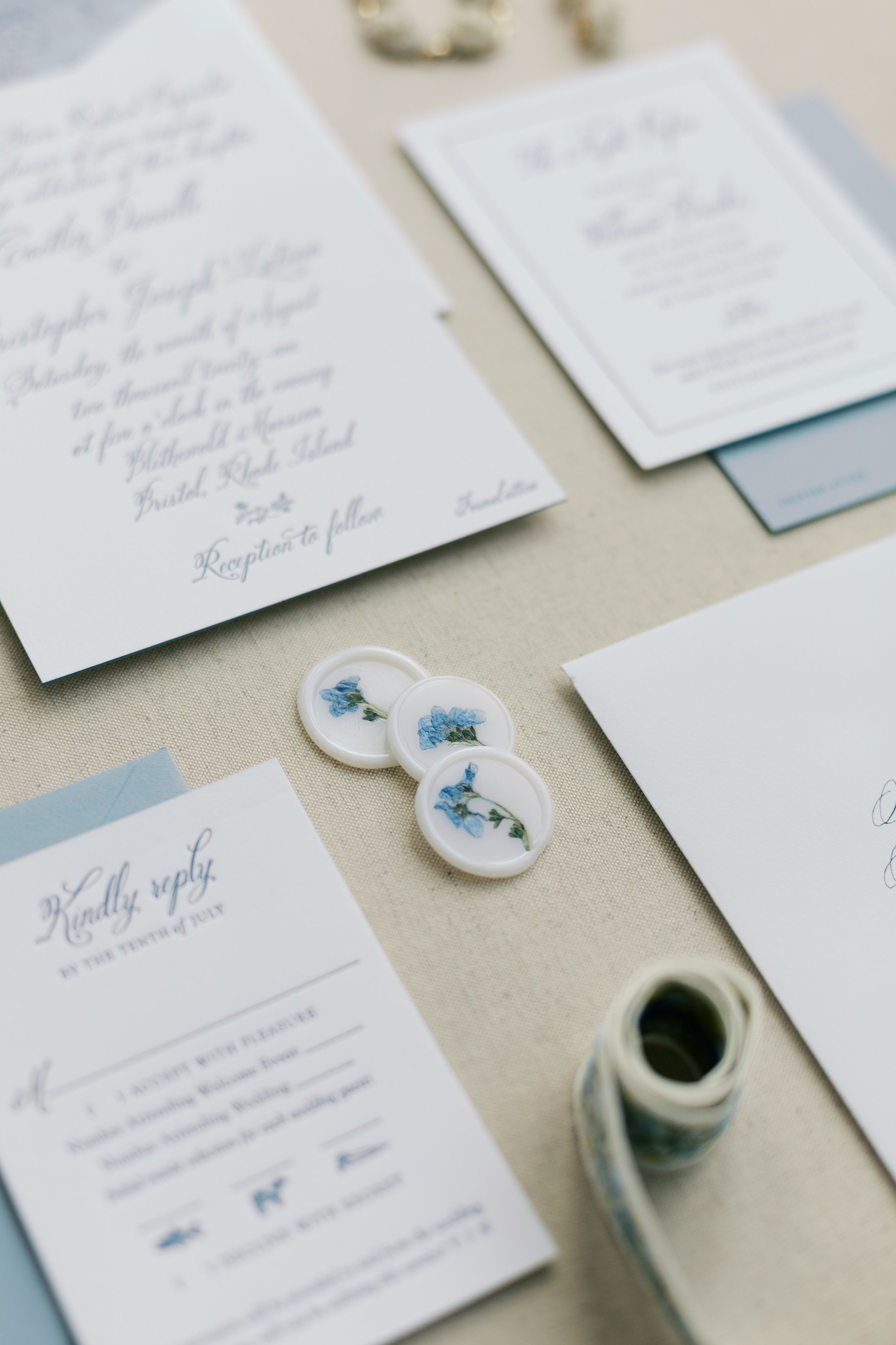 Wedding invitation stationery with blue floral accents, a wax seal stamp, and RSVP card on a beige fabric surface.