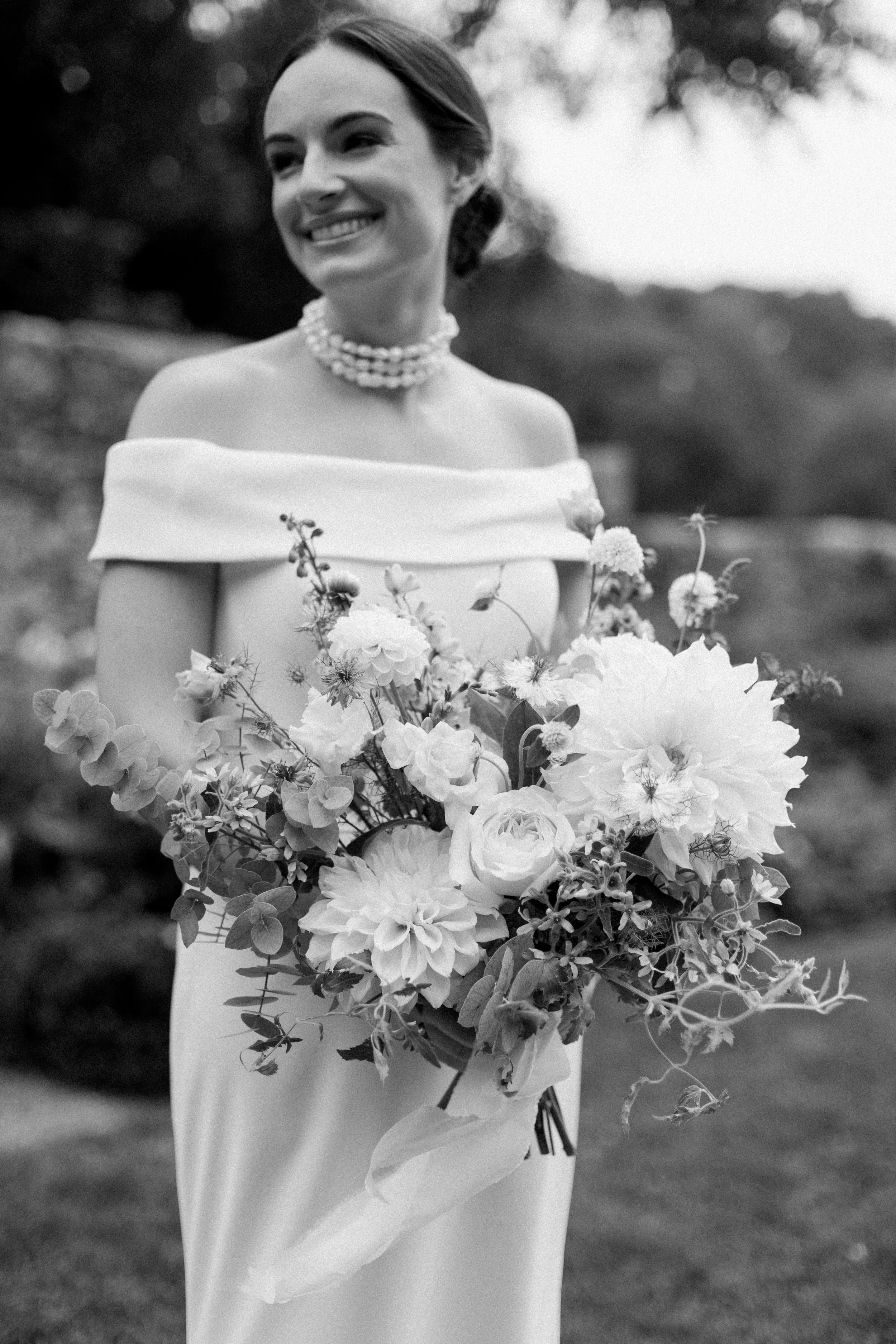 A smiling woman dressed in a wedding gown holding a large bouquet of flowers outdoors.