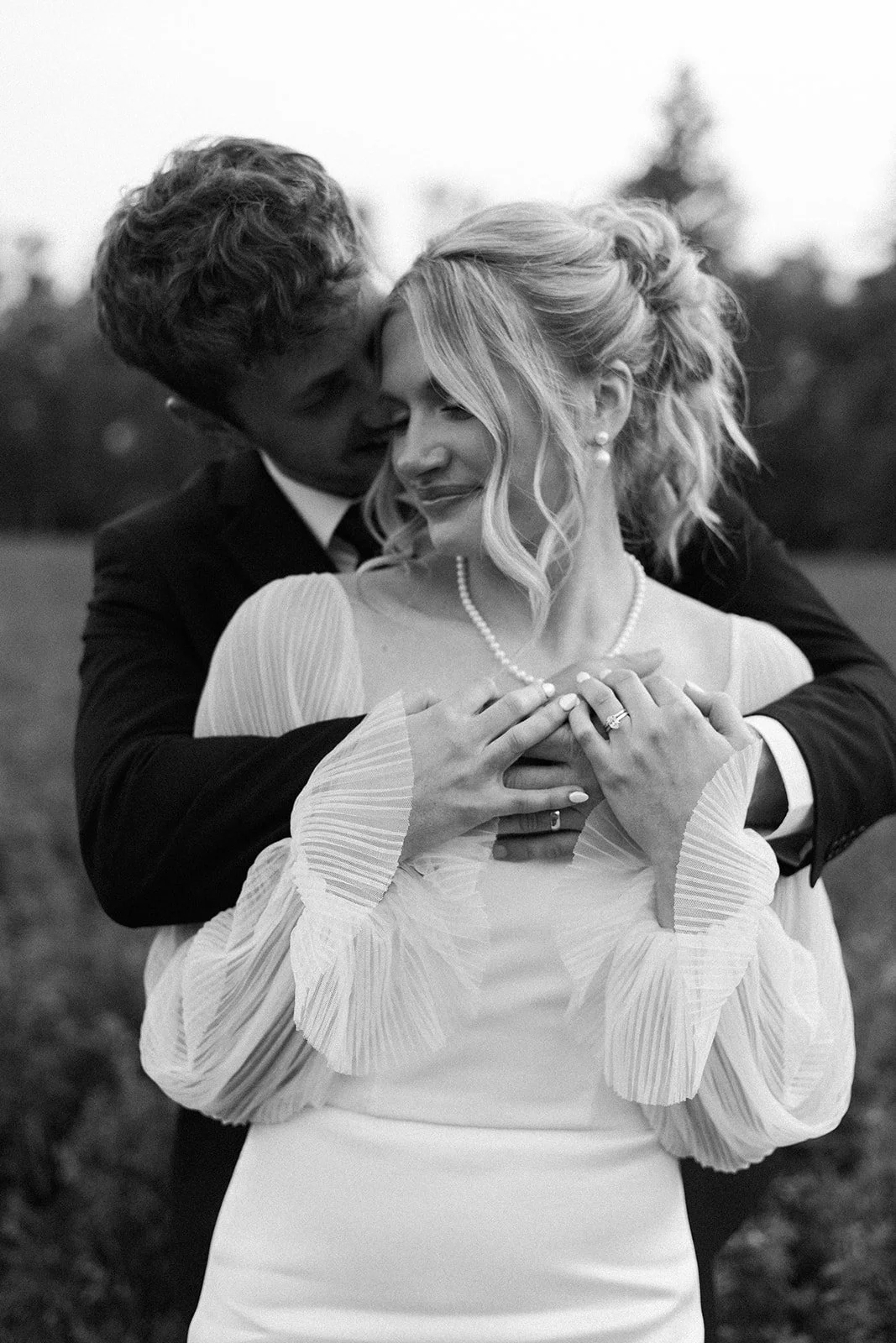 A black and white photograph of a couple in wedding attire embracing outdoors. The man is kissing the woman's temple from behind, and she has her eyes closed with a gentle smile. The woman is wearing a wedding dress with pleated sleeves, pearl earrings, and a pearl necklace. The man is wearing a suit with a tie.