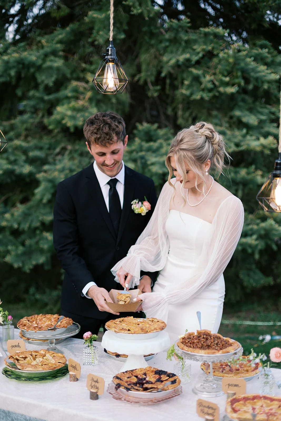 A bride and groom in wedding attire cutting a pie at their outdoor reception. The bride wears a white strapless gown with sheer long sleeves, a pearl necklace, and has blonde hair styled in an elegant updo. The groom wears a black suit, white shirt, 