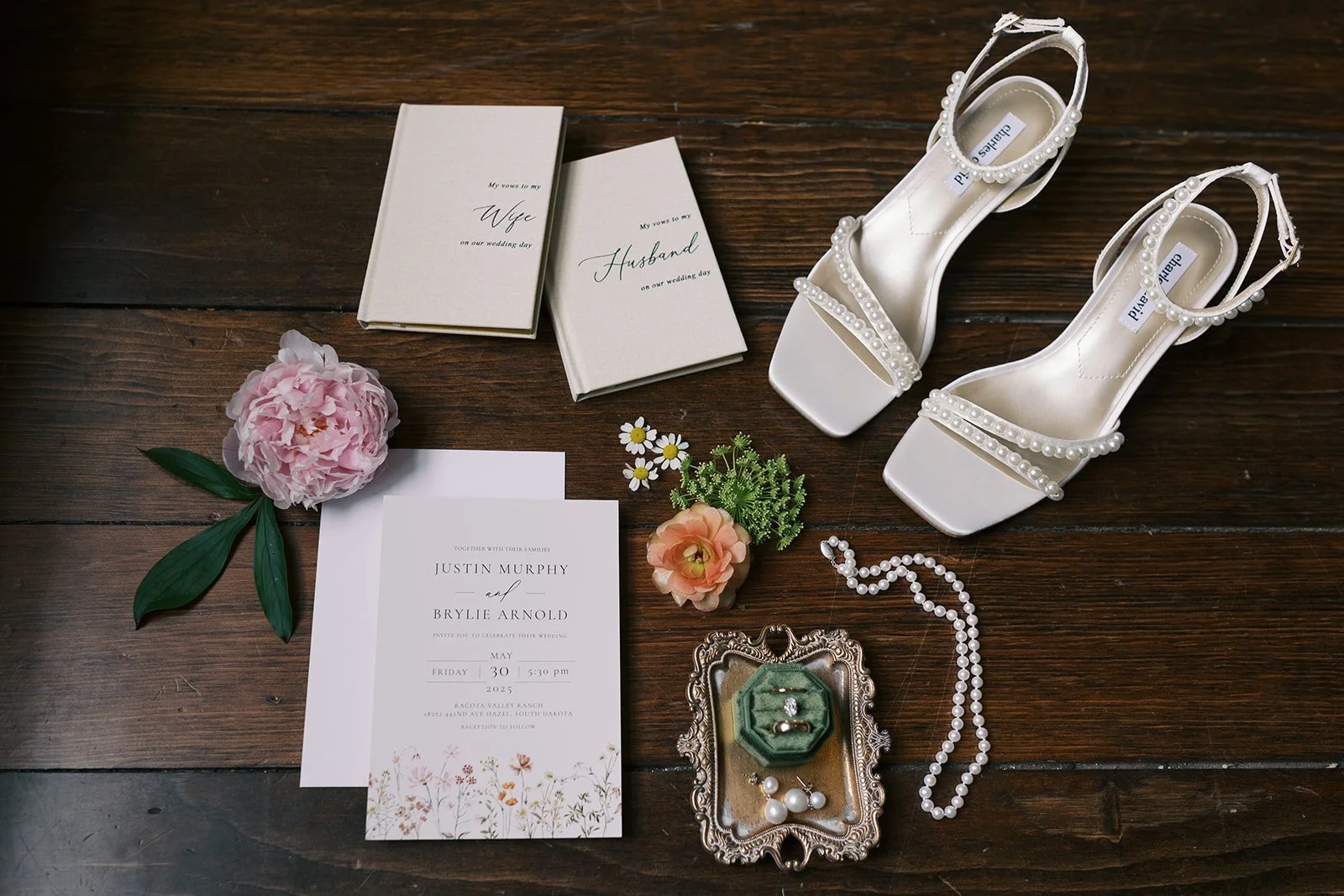 Wedding invitation cards, white pearl-embellished heels, pink peony, small bouquet, pearl necklace, earrings, and a ring in a vintage tray on a wooden surface.