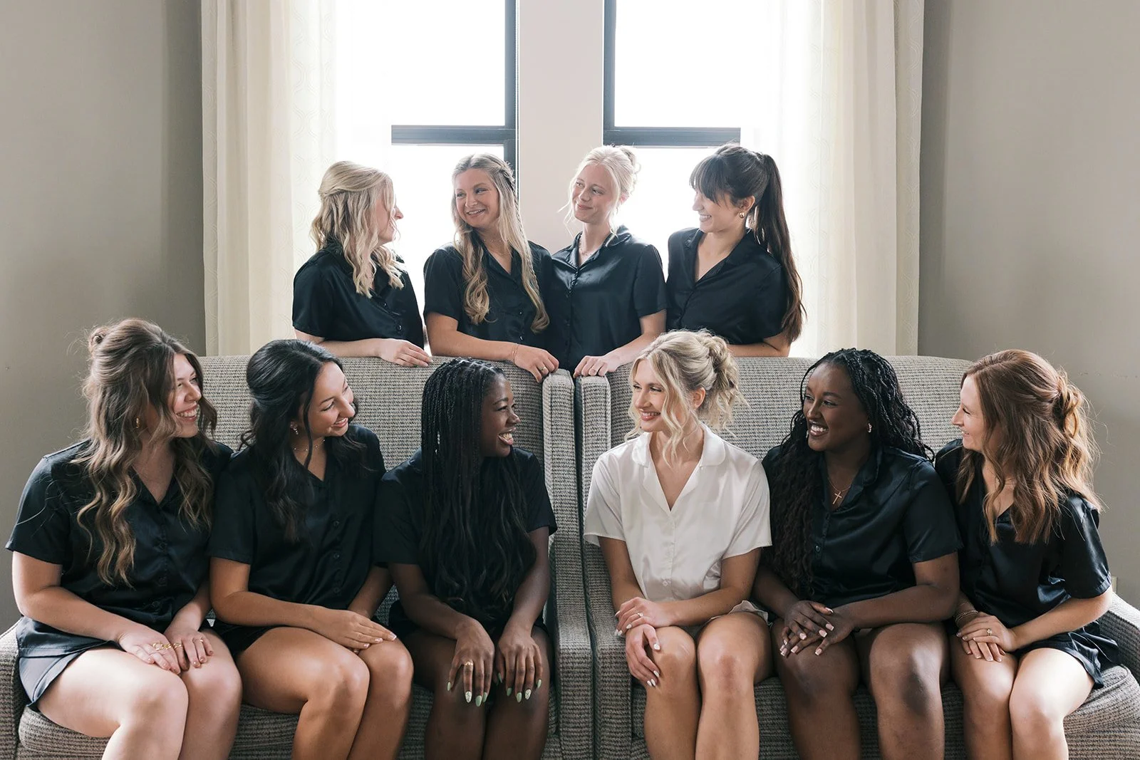 Group of nine women sitting and standing around a couch, smiling and engaging with each other, in a well-lit room with large windows