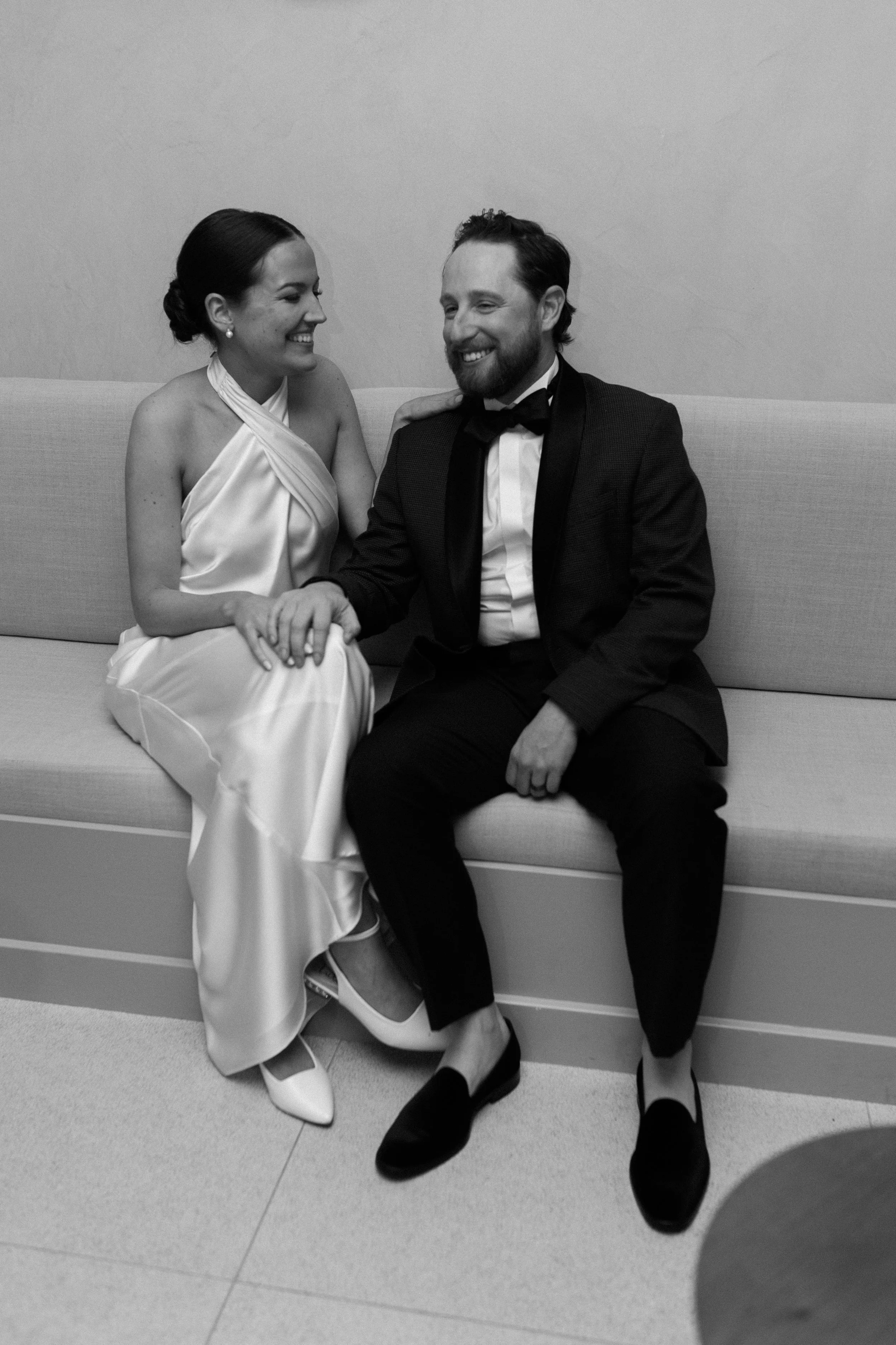 A black and white photo of a smiling woman and man dressed formally, sitting on a sofa, holding hands, and looking at each other.