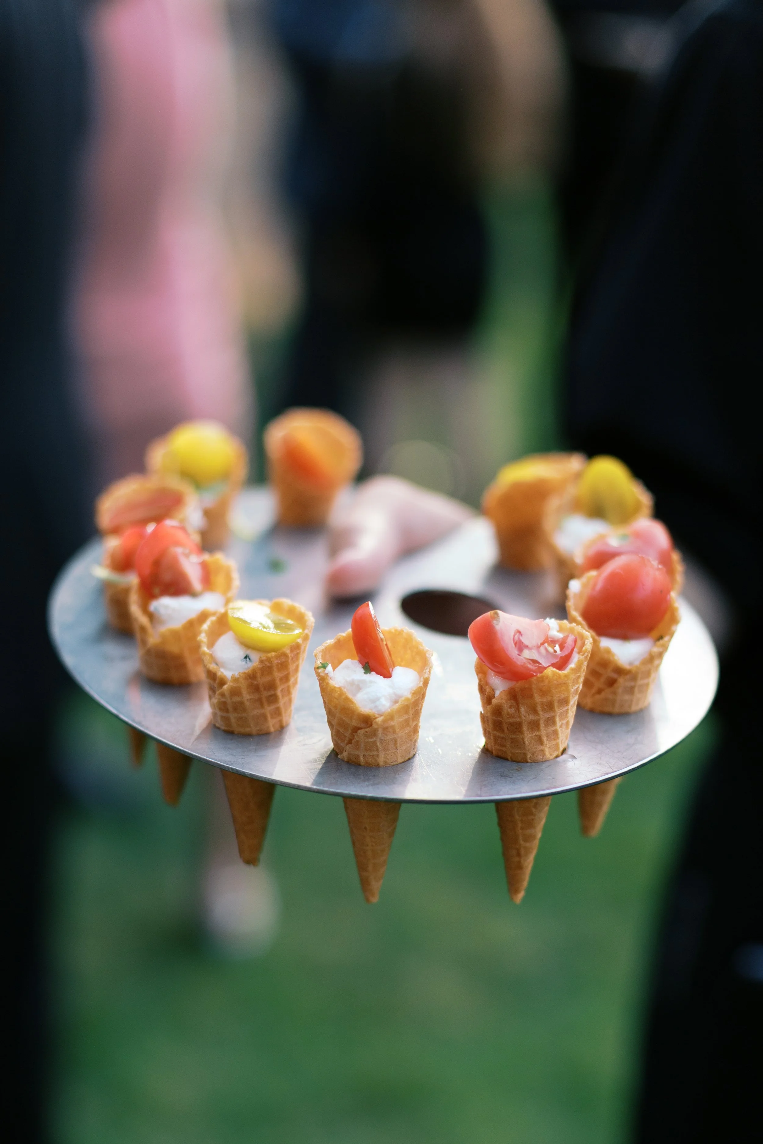 A tray of mini ice cream cone appetizers with colorful toppings and whipped cream, held by a person in a dark shirt, outdoors with a blurred green background.