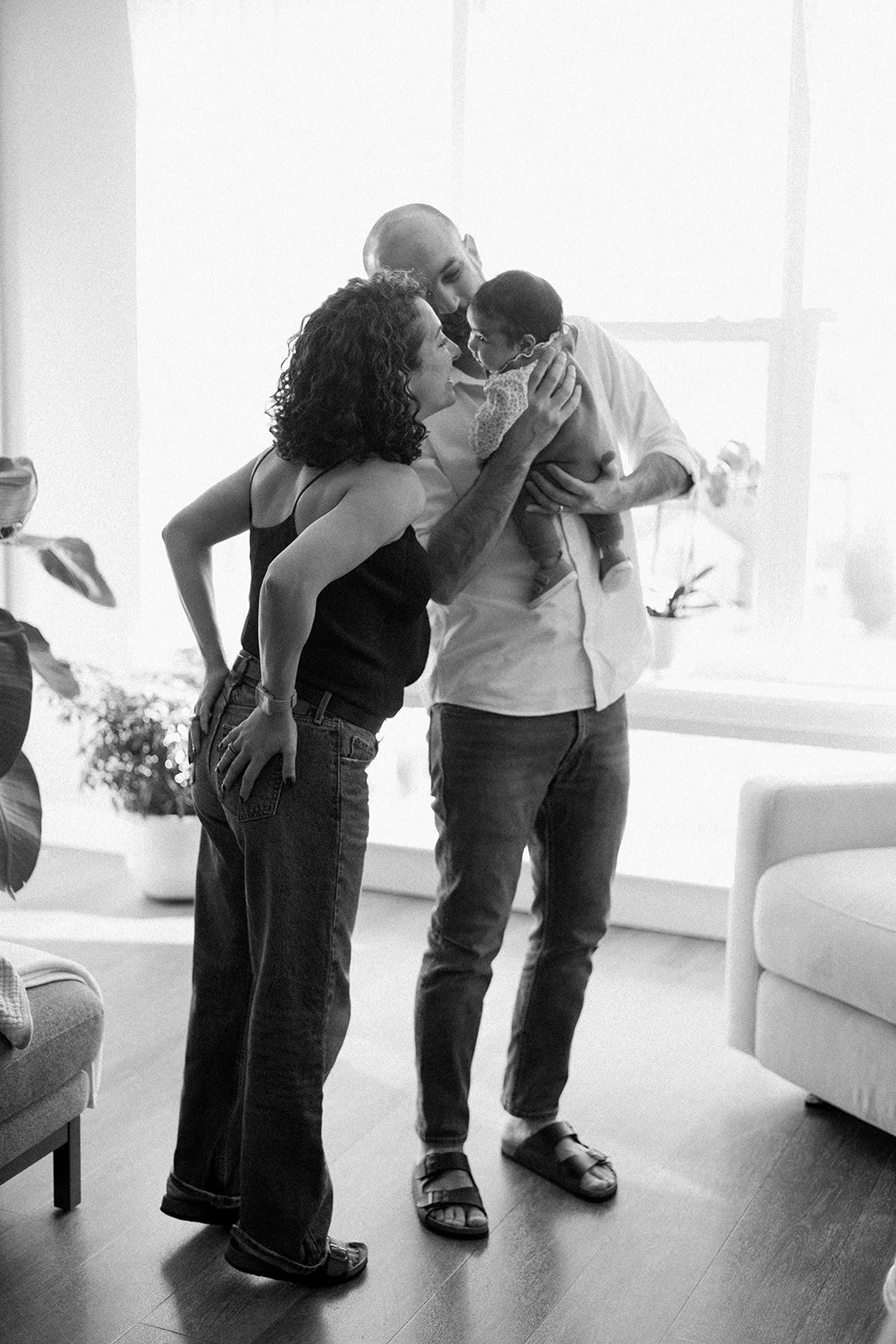 Three people, a man, a woman, and a baby, in an indoor setting. The man is holding the baby, and the woman and man are gazing at the baby with smiles. The scene appears intimate and affectionate.