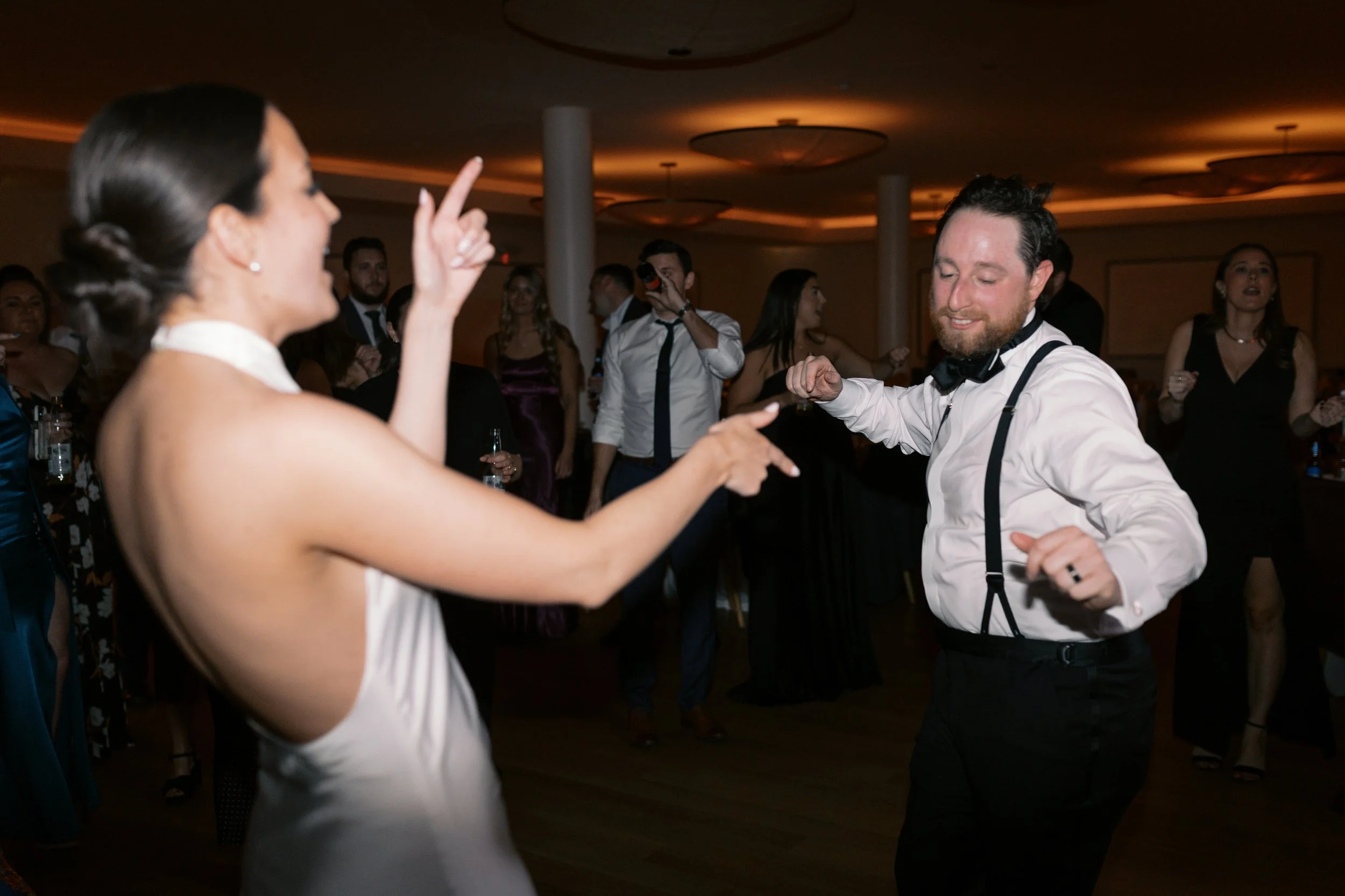 A man and woman dancing at a party or wedding reception, with other guests watching in the background.