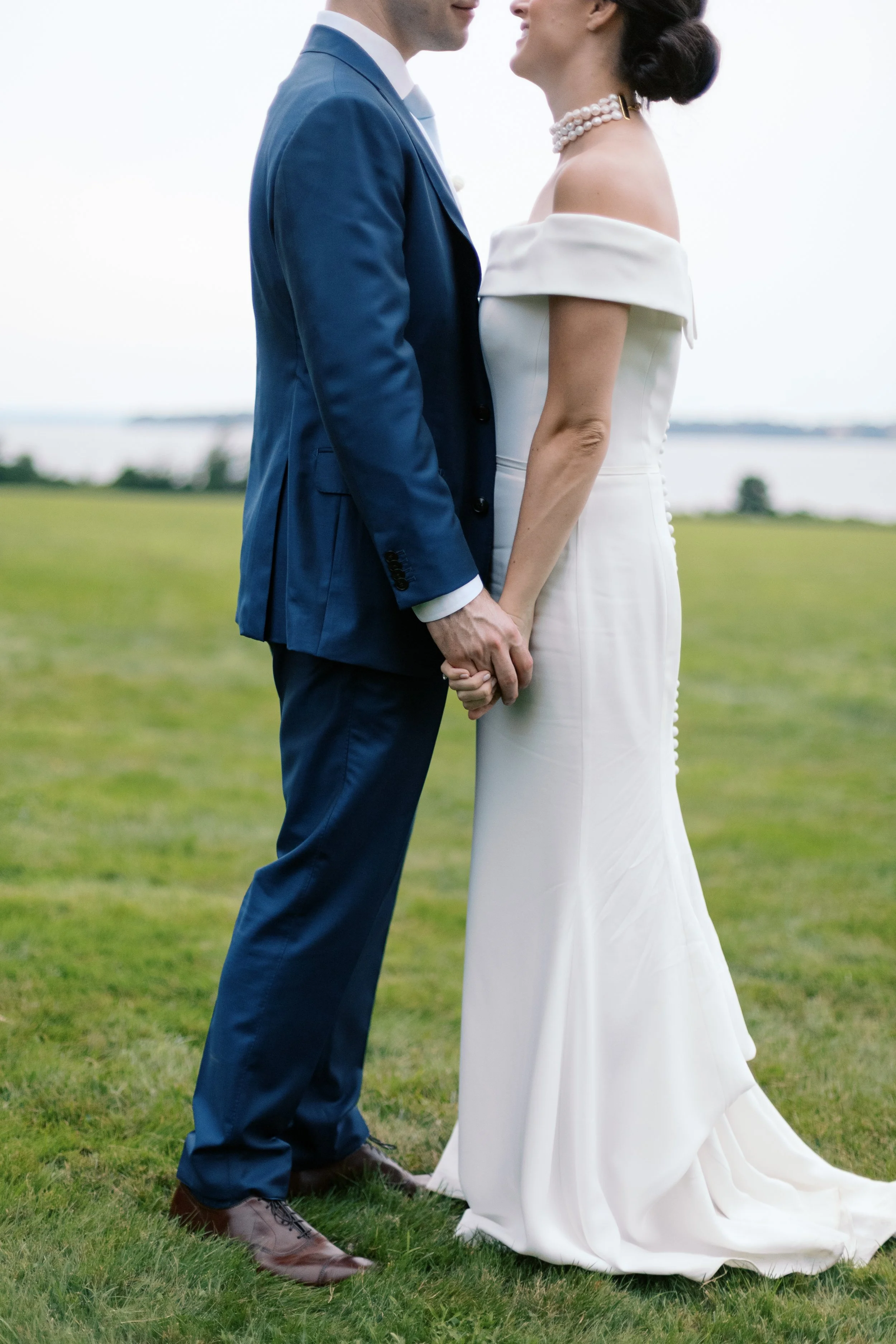 A bride and groom holding hands, standing on grass near a body of water, facing each other during their wedding.