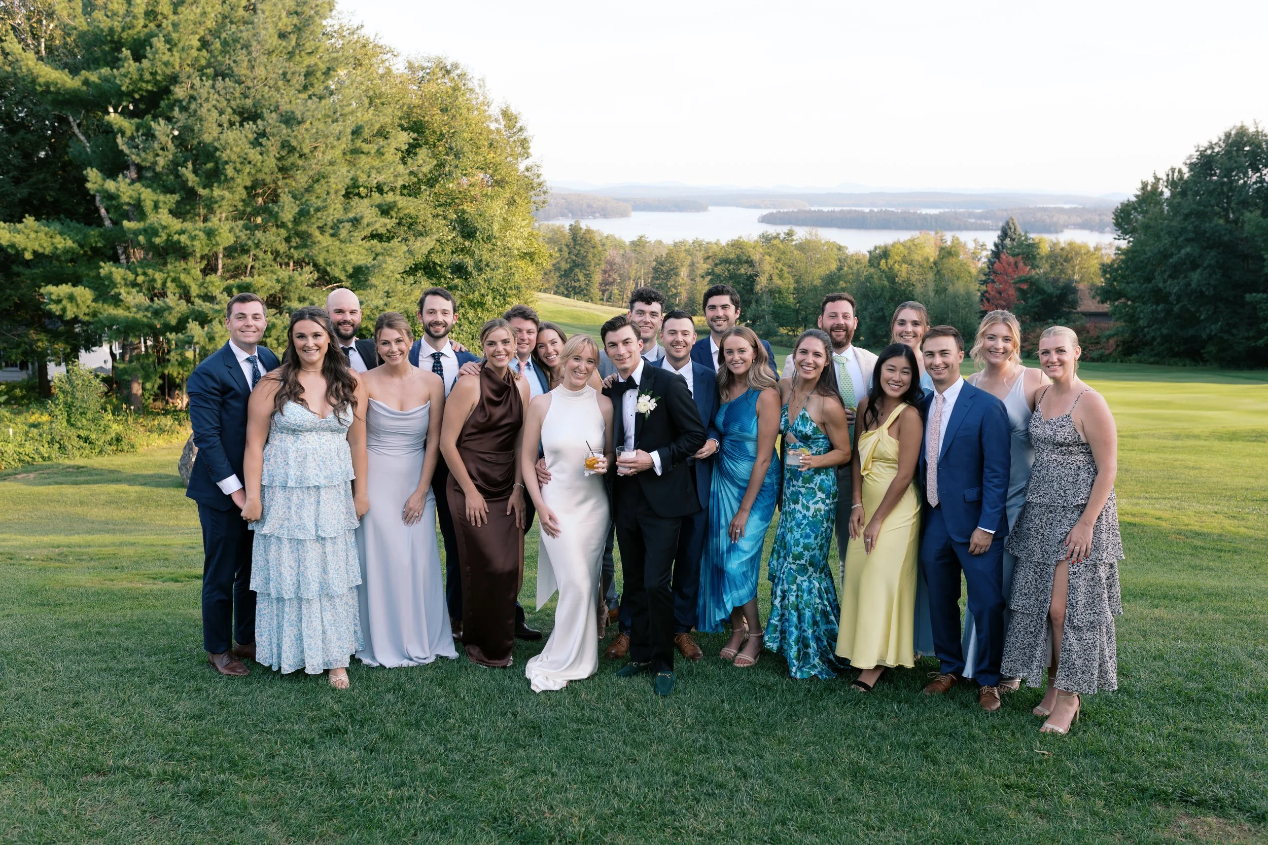 Group of people dressed in formal attire, standing on a lush green lawn with trees and a lake in the background, during daytime.