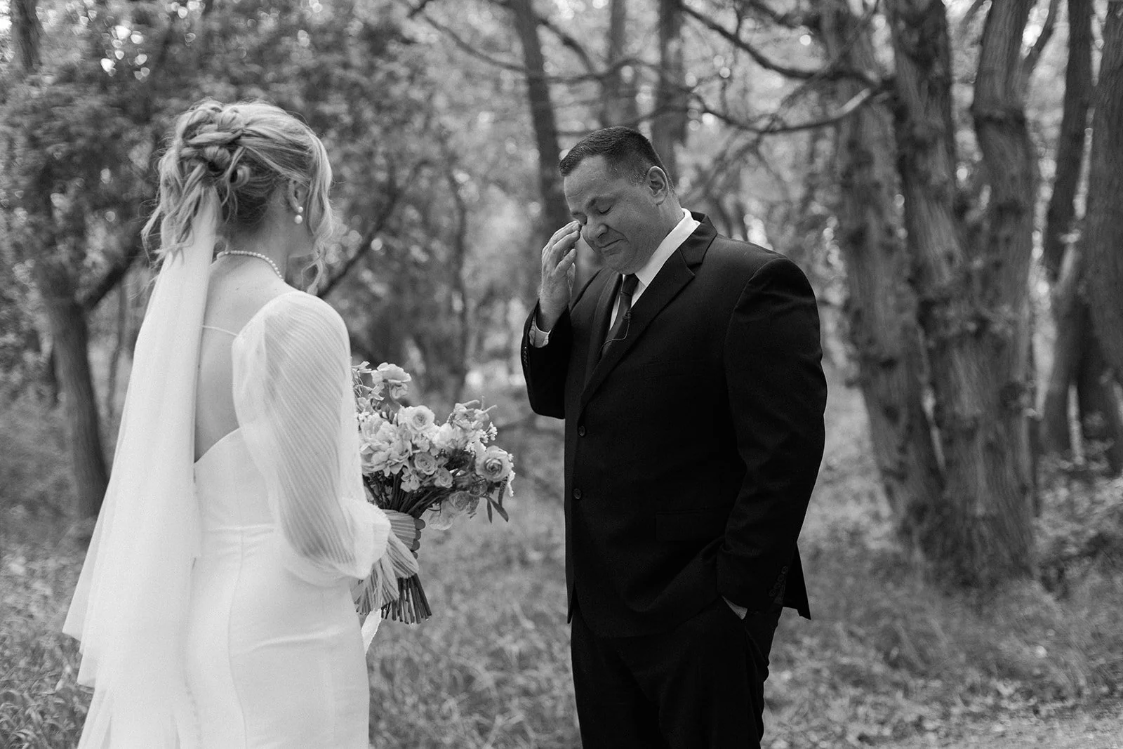 A bride in a white wedding dress holding a bouquet of flowers, standing in front of a groom in a black suit, in an outdoor wooded area.