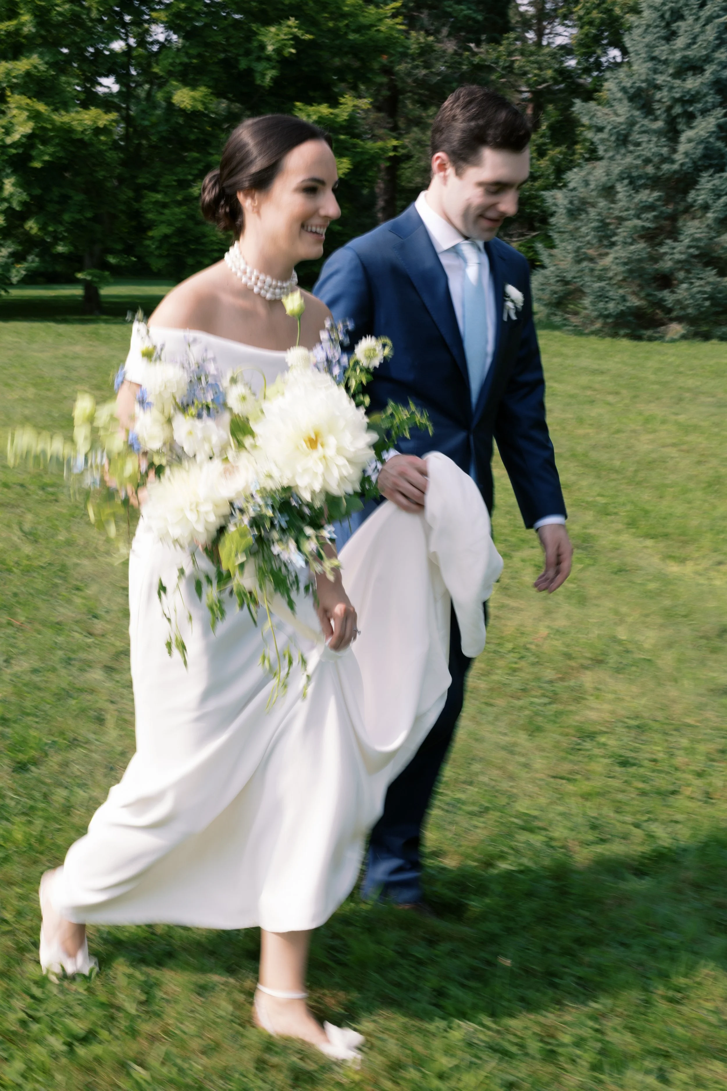 Bride in a white dress holding a bouquet of white and purple flowers, walking with a groom in a dark blue suit on a grassy lawn surrounded by trees.
