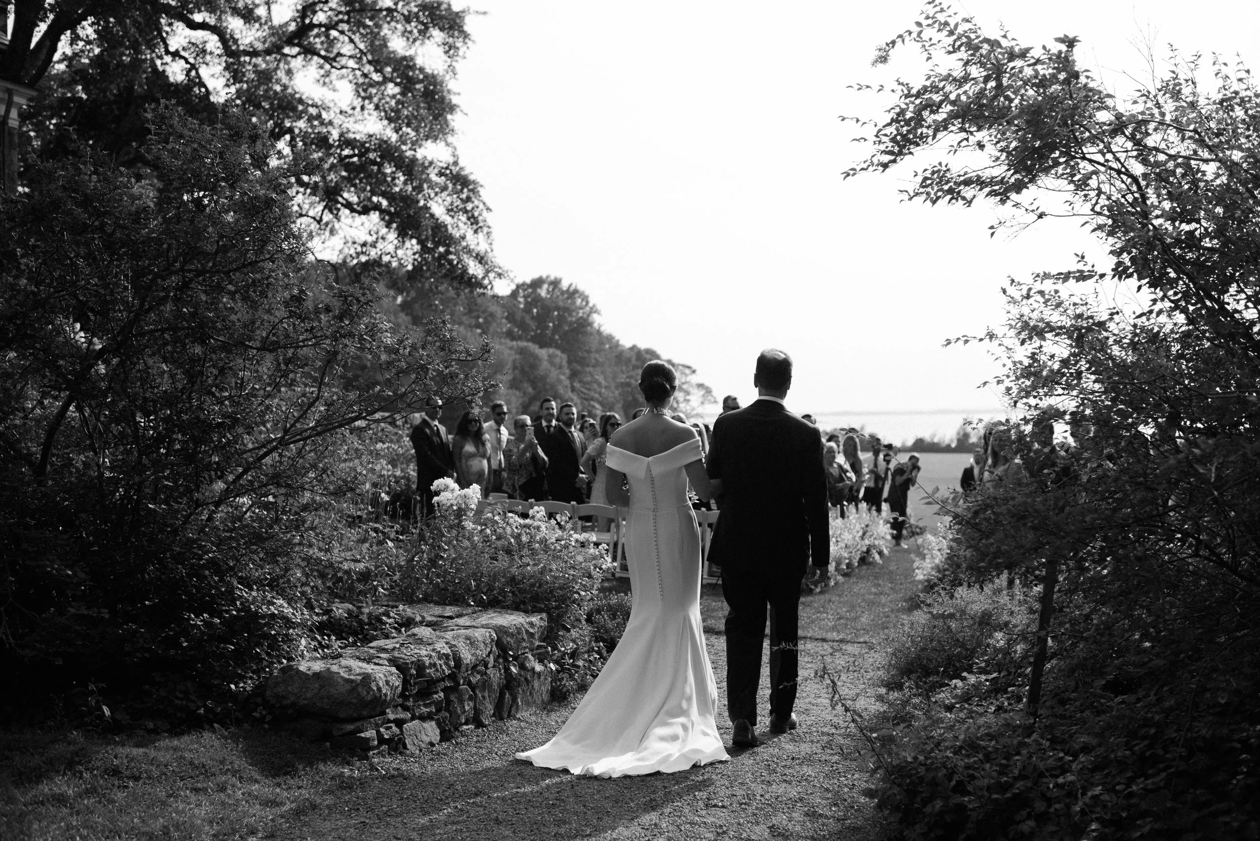 Black and white photo of a bride and groom walking down an outdoor aisle, with guests standing in the background, surrounded by trees and plants.